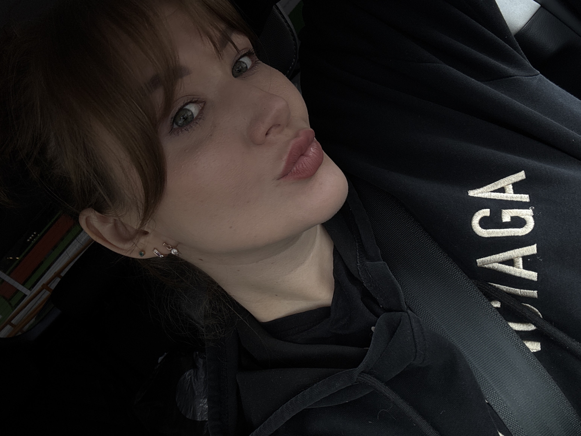 A close-up, selfie-style photograph captures a young woman with light skin and auburn hair, wearing a black hoodie with "BALENCIAGA" embroidered in white on the chest. She is looking towards the camera with her head tilted and puckered lips, her green eyes are visible with makeup. She has multiple piercings in her left ear. A black seatbelt is diagonally across her chest. The background is dark, with some blurred colorful lines visible, possibly from lights or signage. The image does not contain information to determine its location in Chaikovsky, Russia.
