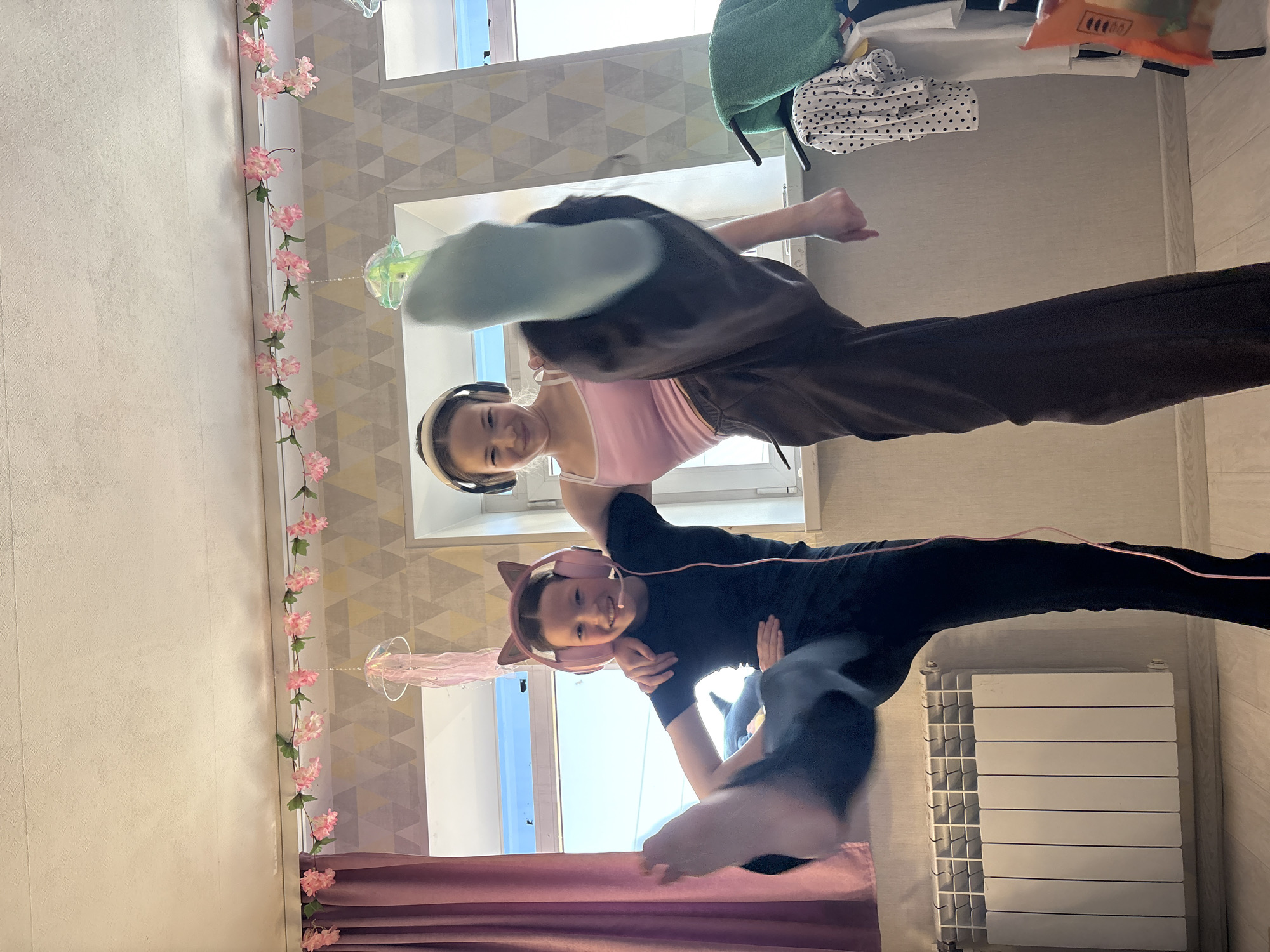 Two girls are posing mid-air in a room decorated with artificial pink cherry blossom garlands. One girl wears a pink tank top and brown sweatpants, headphones, and is smiling. The other girl is wearing a black top and sweatpants, cat-ear headphones, and is also smiling. A radiator is visible in the background. The location is Прикамский, Russia.