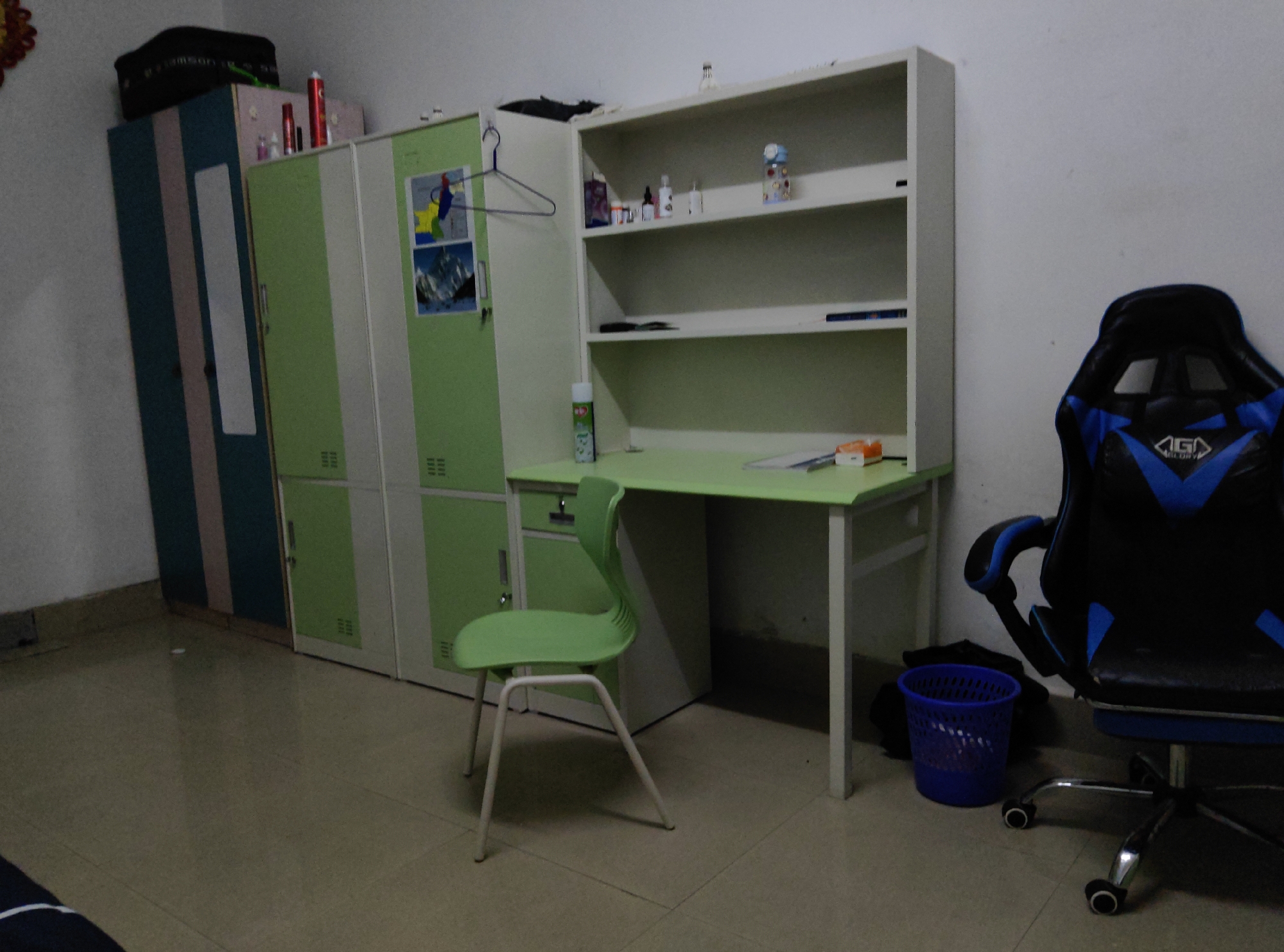 In a room located in the Chongqing High-tech Zone, China, a black and blue gaming chair with the "AGLORY" logo is positioned to the right of a light green desk. The desk has a white frame and a light green top, with a blue mesh trash can next to it. To the left of the desk, a light green chair with a curved back is situated. Behind the desk and chair, a white shelving unit with three shelves is mounted on the wall. The top shelf contains various bottles and a decorative item. The middle shelf is empty, and the bottom shelf has a dark object and a blue pen. To the left of the shelving unit are two tall green cabinets with white accents. The left cabinet has a mirror, and the right cabinet displays two framed pictures. Above these cabinets, a dark bag rests on top of a pink and blue cabinet. Additional items are visible on top of the pink and blue cabinet, including red cans and small bottles. The floor is tiled with a speckled pattern.