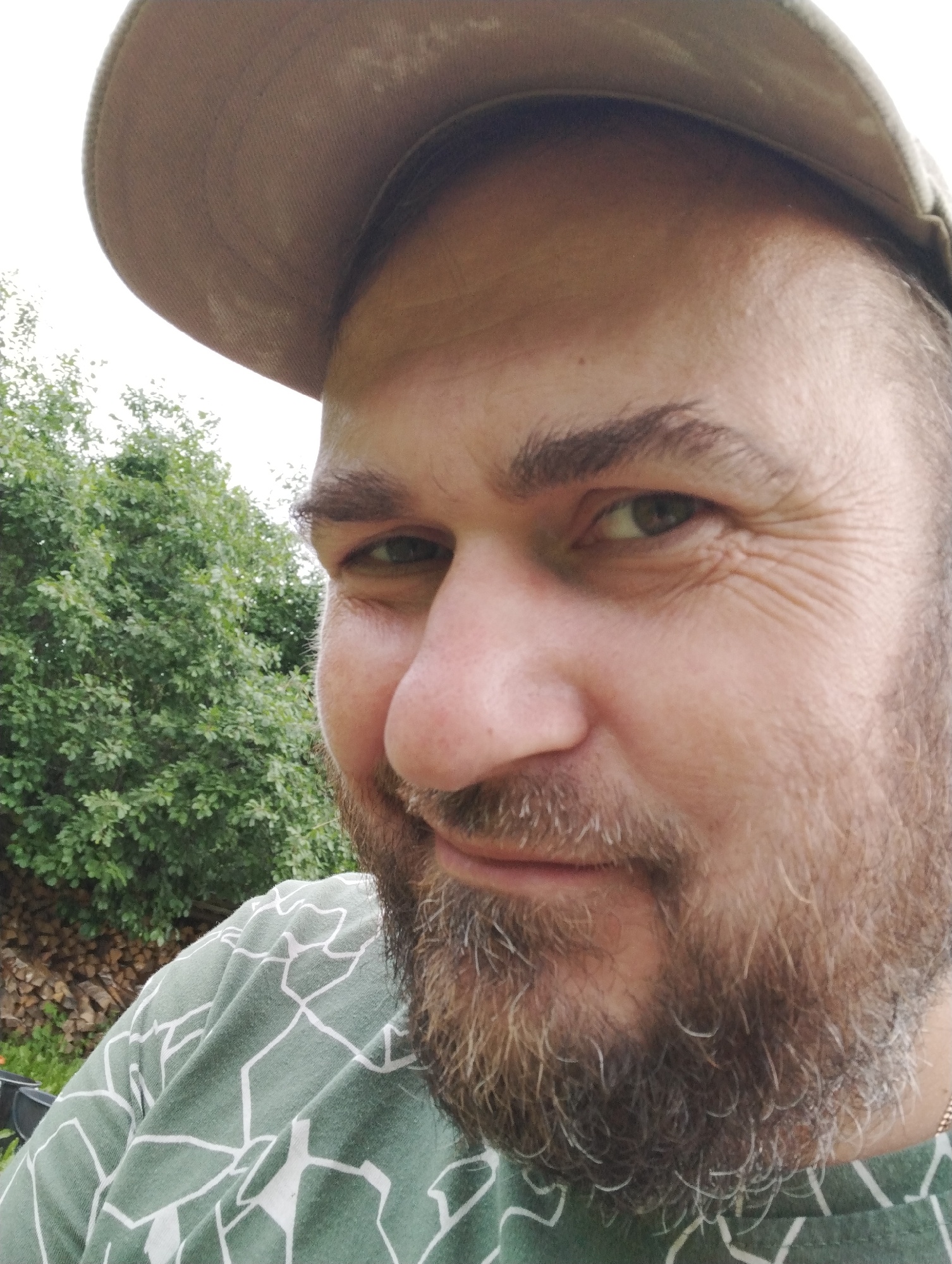 This is a close-up, selfie-style photograph of a man wearing a light brown baseball cap and a green t-shirt with a white abstract pattern. He has a beard and is looking towards the camera with a slight smile. His right eye is visible, and the background consists of lush green trees and a pile of firewood, suggesting an outdoor setting. The lighting indicates it is daytime, likely a sunny or overcast day, as there are no harsh shadows. The image seems to have been taken in a rural or natural environment. The location context provided is Ширяево (ильинский), Russia. There is no visible text in the image.