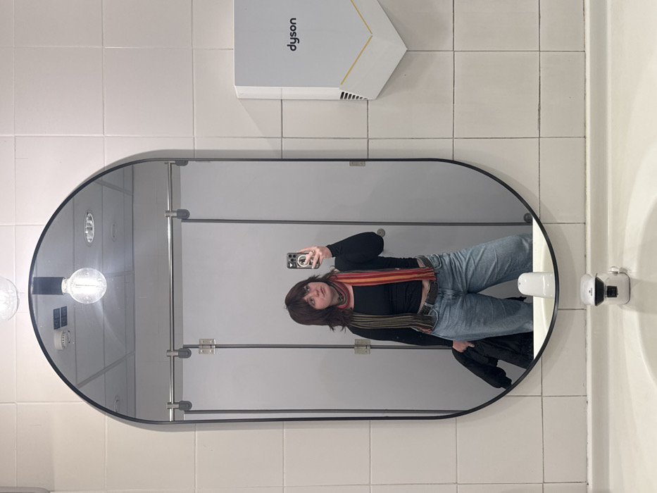 The image depicts a female individual taking a mirror selfie in a public restroom. The exact location within Sesto Fiorentino, Italy, is not identifiable from the visual evidence.
The central subject is a young adult female, reflected from the waist up in a large, vertically oriented oval mirror with a black frame. She has medium-length brown hair with bangs and is wearing a long-sleeved black top, light blue loose-fitting jeans, a dark belt, and a multi-colored striped scarf with red, orange, green, and brown hues draped around her neck. In her raised right hand, she holds a light-colored smartphone with a clear, MagSafe-compatible case, positioned to capture her reflection. Her left hand holds a folded dark-colored jacket or coat. Her gaze is directed towards the smartphone/mirror.
The background, visible both directly and in the mirror's reflection, consists of walls clad in large, rectangular white tiles with light grey grout. To the right of the mirror, a white Dyson brand hand dryer with a gold or tan base is mounted on the tiled wall. Below the mirror, a white ceramic sink basin is visible, equipped with a white, modern-style faucet featuring a black top. Reflected in the mirror are two light-grey or white public restroom stall doors with silver horizontal rails and a central hinge. The ceiling, also reflected, appears to be a light-colored grid ceiling with several embedded circular lights and a smoke detector. A bare incandescent light bulb in a black fixture is suspended from the ceiling, visible above the mirror's top edge. The lighting is bright and appears to be artificial overhead illumination.
