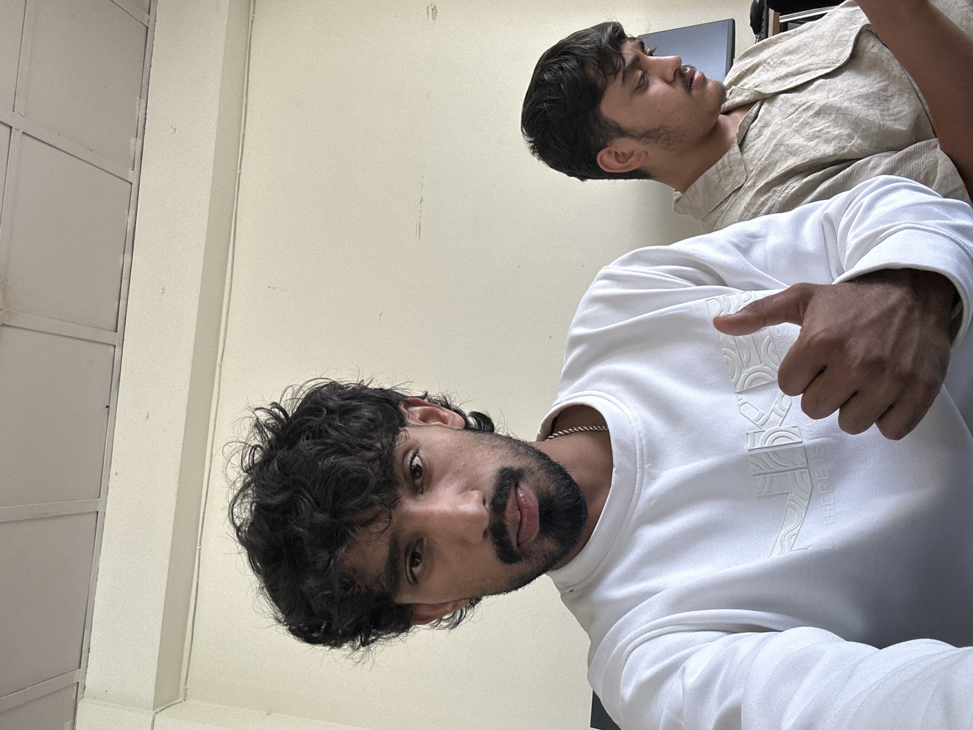 Two young men are positioned in a brightly lit room with plain, cream-colored walls and a white ceiling grid. The man in the foreground, on the left, is closer to the camera and looking directly at it. He has curly dark hair, a beard, and is wearing a white sweatshirt with a textured design and the words "THERE IS NO" visible. He is giving a thumbs-up gesture with his right hand. In the background, slightly to the right and further from the camera, is another young man with dark hair who is looking down at something in his hands, possibly a phone. He is wearing a light-colored, patterned short-sleeved shirt. A computer monitor is visible behind him. The scene appears to be indoors, possibly an office or study space.