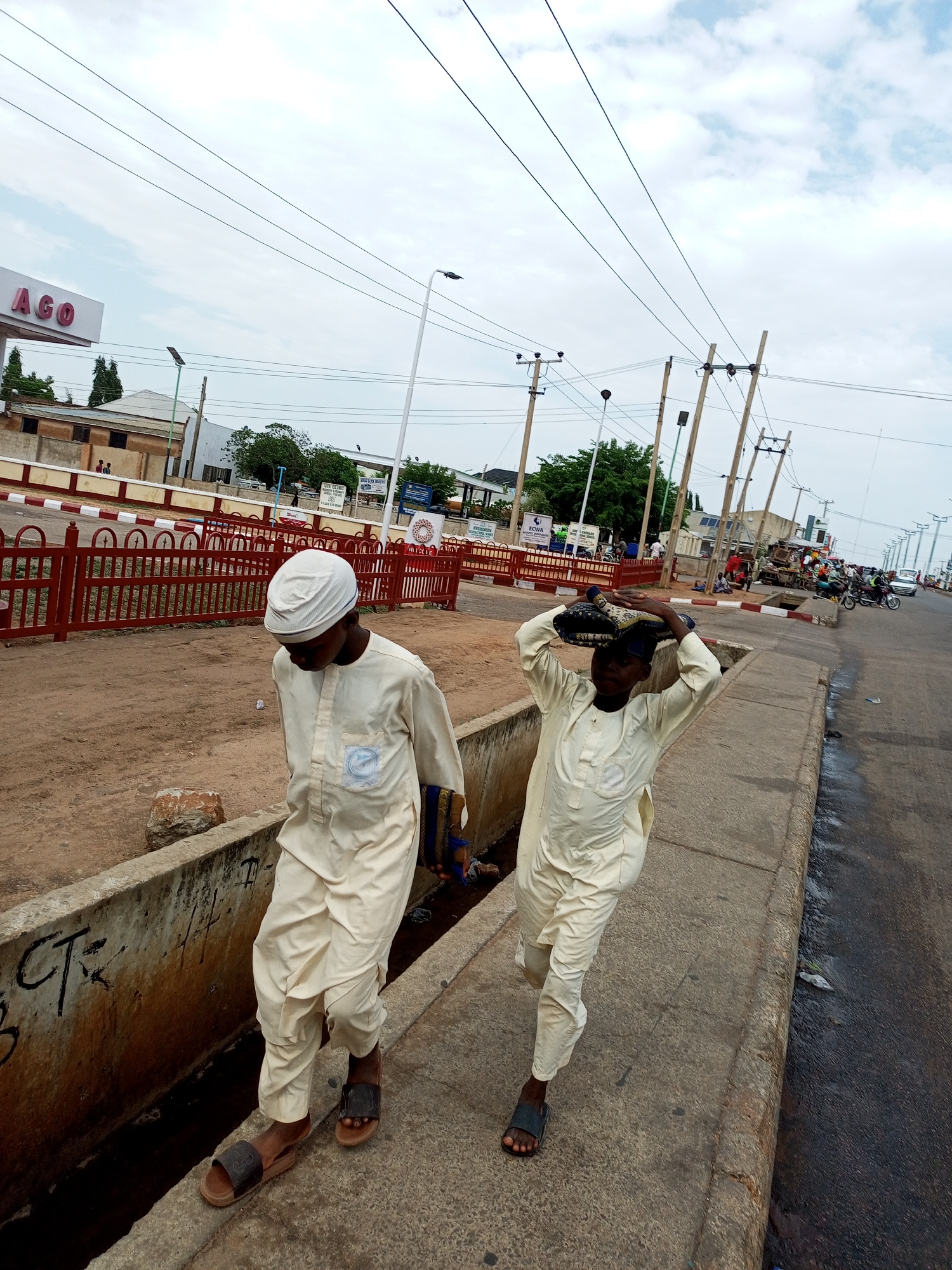 Two young boys, likely of Hausa ethnicity, walk along a sidewalk in Gombe, Nigeria. The boy on the left, wearing a white turban and a flowing cream-colored robe, carries a rolled-up dark blue and gold fabric bundle in his right hand. The boy on the right, also in a cream-colored robe and a patterned cap, balances a similar bundle on his head with both hands. Both boys wear simple slip-on sandals. They are walking on a concrete sidewalk adjacent to a dirt path and a drainage ditch. The background reveals a street scene with a red metal fence, buildings with signs, utility poles with electrical wires, and several parked motorcycles and vehicles, indicating a populated urban area. The sky is overcast. The image captures a moment of daily life in Gombe.