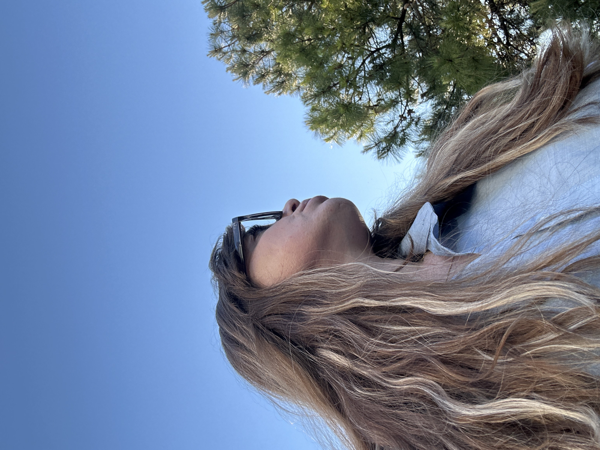 A low-angle photograph depicts a person with long, highlighted brown hair wearing glasses, looking upwards towards the sky and a tree. The sky is clear and blue. The tree's foliage is visible in the upper portion of the frame. No specific information about the location in Batumi, Georgia is discernible from the image.