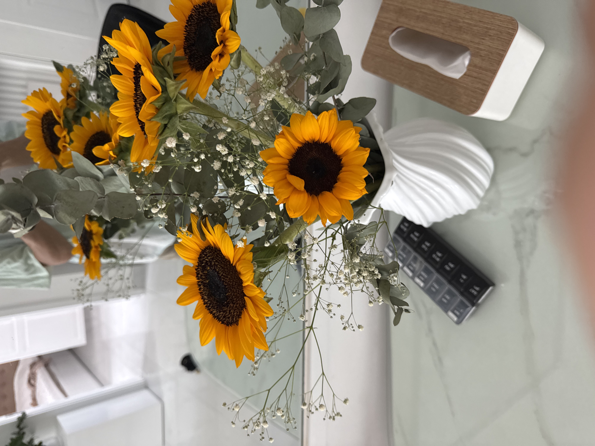 This image captures a close-up of a bouquet of sunflowers in a white vase, placed on a reflective surface, likely a table with a marble-like pattern. The sunflowers are vibrant orange with dark centers, interspersed with delicate white baby's breath and eucalyptus leaves. In the foreground, to the left of the vase, is a black pill organizer with multiple compartments. To the right of the vase is a wooden and white tissue box. The background, visible in the reflection, suggests an indoor setting with a bed and possibly a closet. The overall scene conveys a sense of domestic tranquility. There is no specific indication of the location being in Bac Nha Trang Ward, Vietnam.