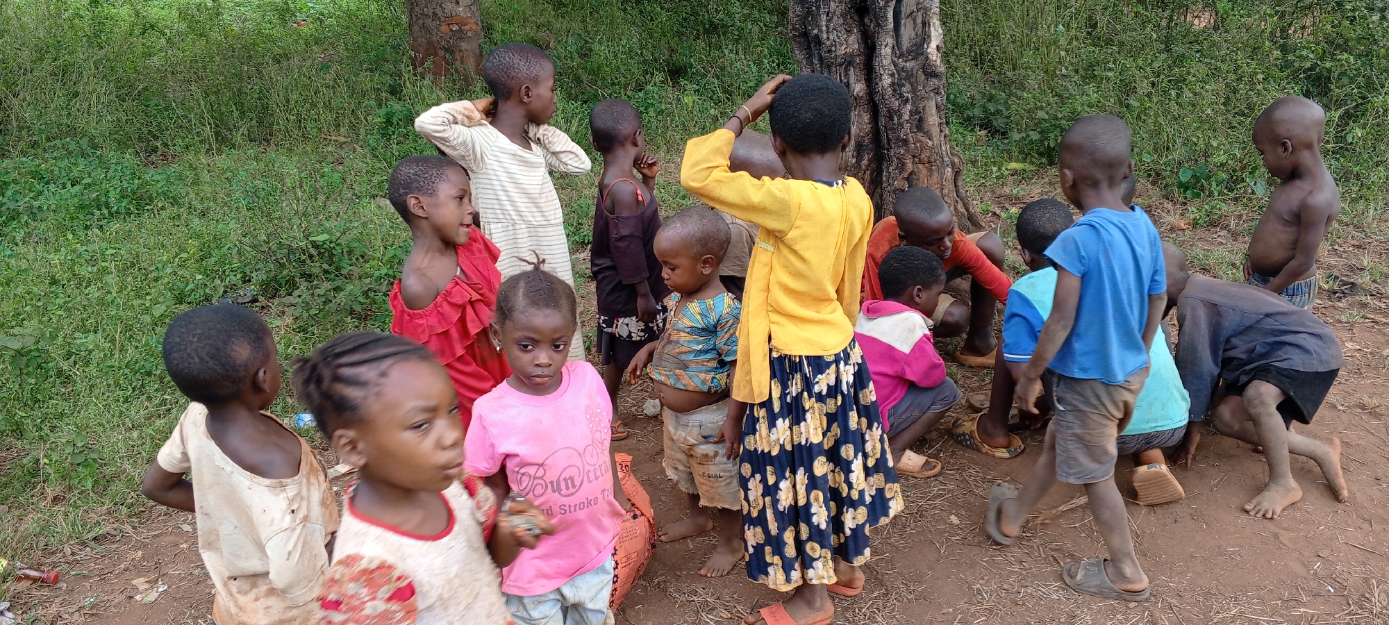 The image shows a group of approximately 15 children gathered in an outdoor area, likely in Ngalula, Democratic Republic of the Congo. The environment is characterized by a dirt ground, some short green vegetation, and a tree trunk visible in the background. The children are of varying ages and are dressed in assorted clothing, including t-shirts, dresses, and shorts, some of which appear worn. Some are barefoot, while others wear sandals. The children are engaged in various activities; some are standing, some are looking at the camera, while others seem to be playing near the tree trunk. Their skin is dusty. There is an orange bag on the ground in front of the child in the pink shirt.