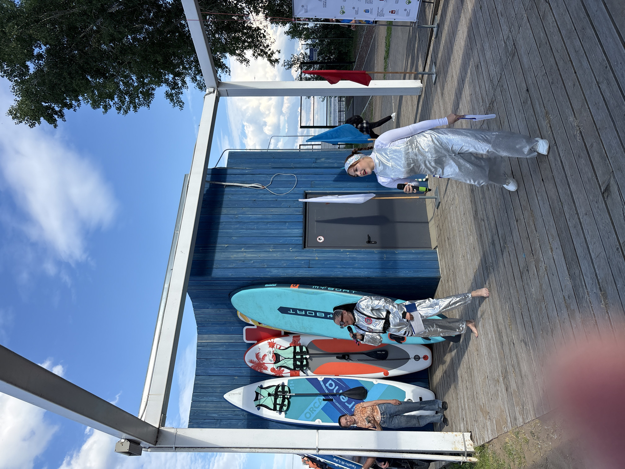 In Chaikovsky, Russia, a group of people are gathered on a wooden deck next to a blue wooden building. The scene appears to be an outdoor event, possibly a festival or competition, given the presence of several paddleboards leaning against the building. The sky is clear and blue with some scattered clouds, suggesting a pleasant day, likely daytime.
Two individuals are prominently featured. One person, dressed in a shimmering silver outfit and a white long-sleeved shirt, is holding a microphone and a piece of paper, appearing to be addressing the crowd or participants. They are wearing a white headband. Another person is lying on one of the paddleboards, wearing a patterned shirt and jeans, and seems to be observing the proceedings. A third person, also in a silver outfit, is lying on a paddleboard with their feet extended.
The paddleboards are colorful and varied. One is light blue with "MYBOAT" printed on it. Another features a tropical design with palm trees. The third paddleboard is predominantly blue and white with "ORCA" and "Rena" visible.
In the background, a red flag is visible on a pole, and banners with text are hung above. The overall atmosphere seems relaxed and festive, with the clear sky and outdoor setting contributing to a summery feel. The presence of multiple paddleboards suggests a water-related activity is part of the event.