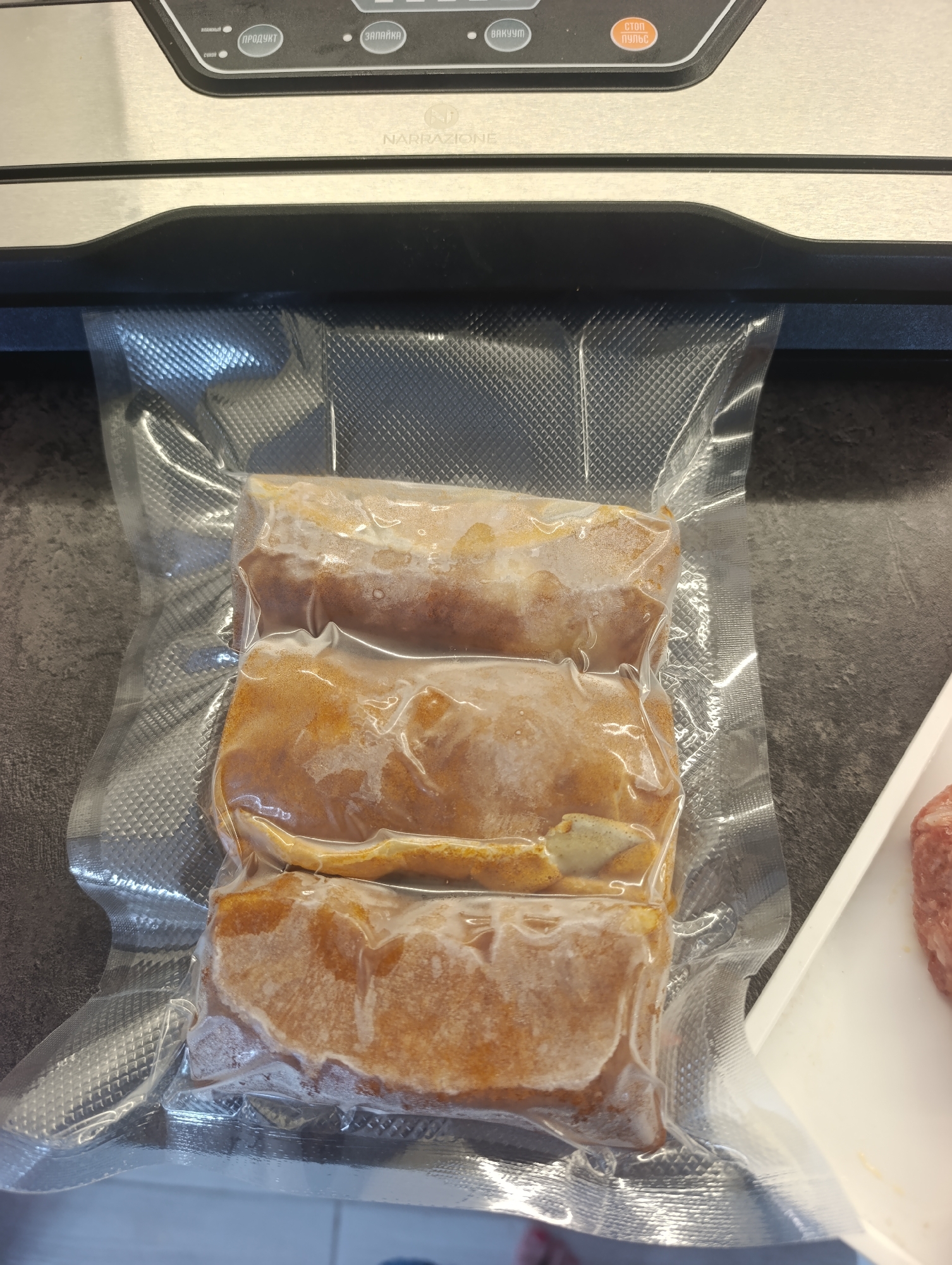 Three frozen, breaded meat patties are vacuum-sealed in a clear plastic bag. The bag is placed on a dark gray countertop, with the top portion of a vacuum sealer machine visible above the bag. The vacuum sealer has Russian text labels on its control panel, including "ПРОДУКТ" (Product) and "ЗАПАЙКА" (Sealing). To the right of the bag, a white tray with a partially visible raw meat patty is present. The scene appears to be in a kitchen environment.