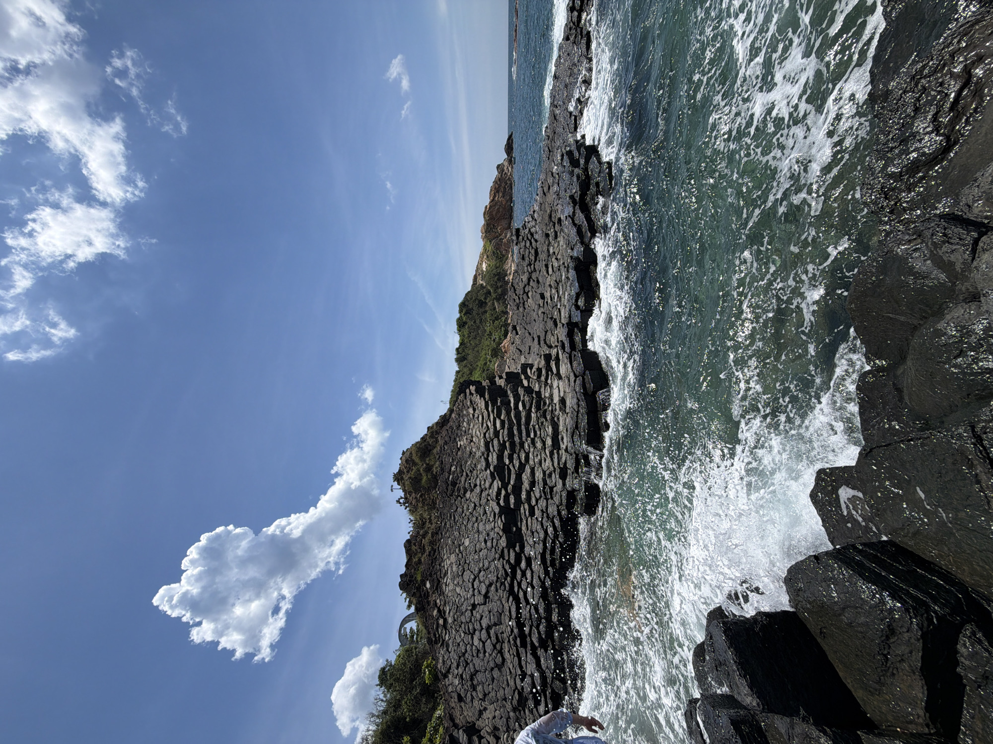 A low-angle, wide shot captures the columnar basalt formations of Gành Đá Đĩa in Phú Lương, Vietnam, with the turquoise sea and a bright blue sky. In the foreground, dark, wet basalt rocks are partially submerged as waves crash against them, creating white foam and spray. A person in a white shirt and blue pants stands near the left edge of the frame, facing away from the camera and towards the water. The midground features a large, stepped formation of black columnar basalt, rising to a vegetated cliff. The background shows more of the rocky coastline, lush green trees, and the horizon where the sea meets the sky. Scattered white clouds dot the bright blue sky, with one particularly large, billowy cloud prominently positioned above the basalt formations. The overall scene conveys a natural, coastal landscape bathed in sunlight.