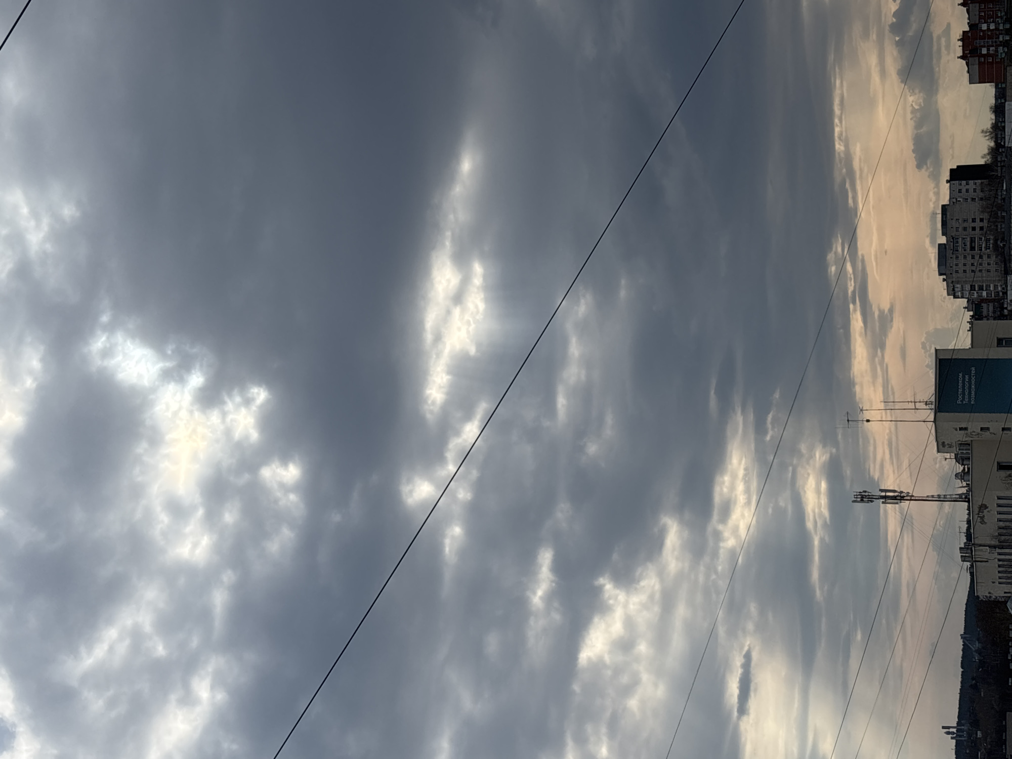 The image shows a view of the sky over the city of Chaikovsky, Russia. The sky is predominantly covered with gray and white clouds, with some areas appearing darker and more ominous. Glimmers of light shine through breaks in the clouds.
In the lower portion of the frame, there is a cityscape consisting of buildings and structures, including residential buildings. Utility poles and wires are visible extending across the sky. The perspective is from a low angle, looking upward towards the sky. The building on the right has a sign with the name of Rostelecom company.