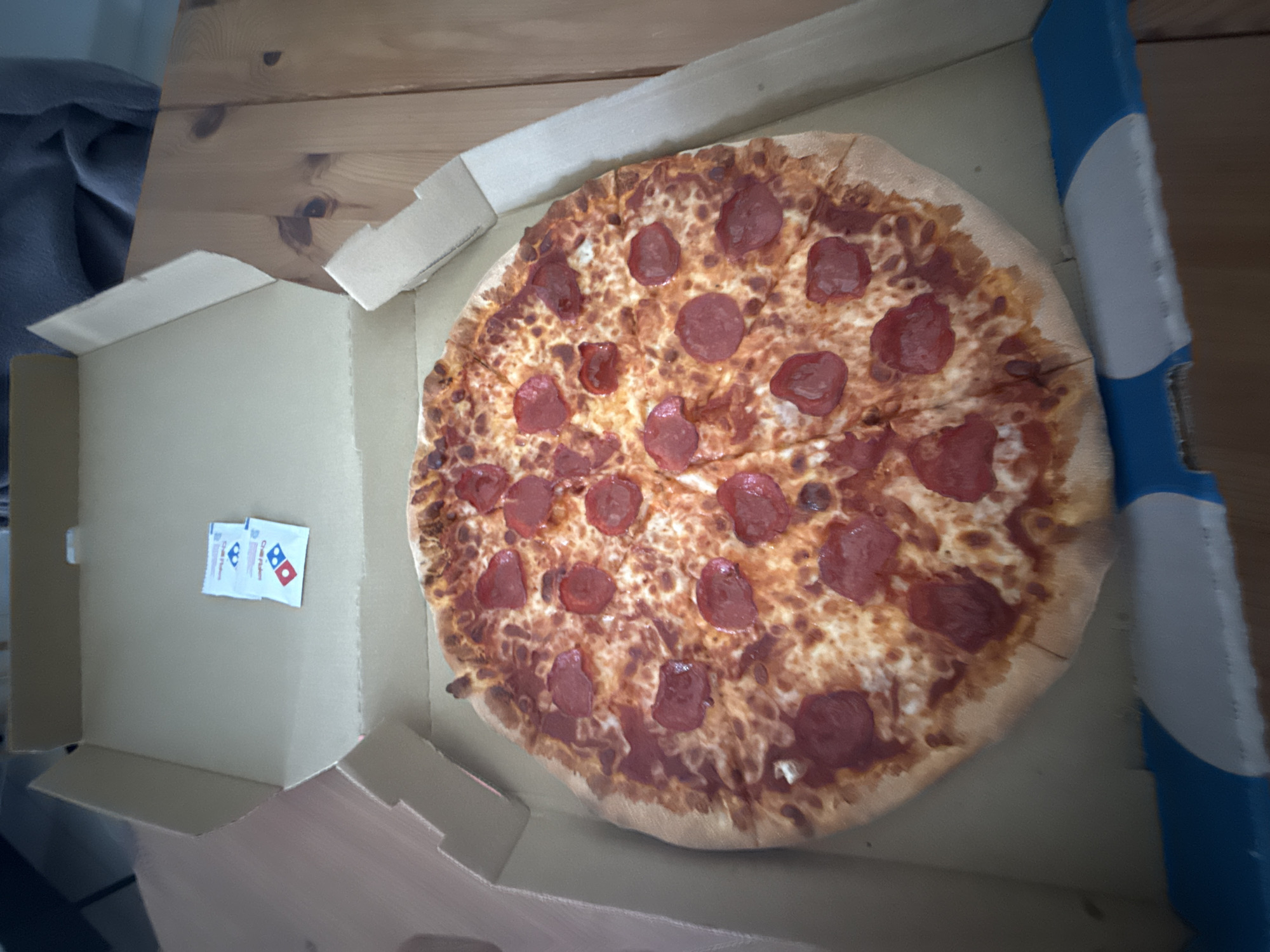 A pepperoni pizza sits in an open Domino's pizza box on a wooden surface. Two small packets, possibly seasoning or parmesan cheese, are in the box next to the pizza. The box has blue accents. The location of this scene in Kuala Lumpur, Malaysia is not discernible from the given image.