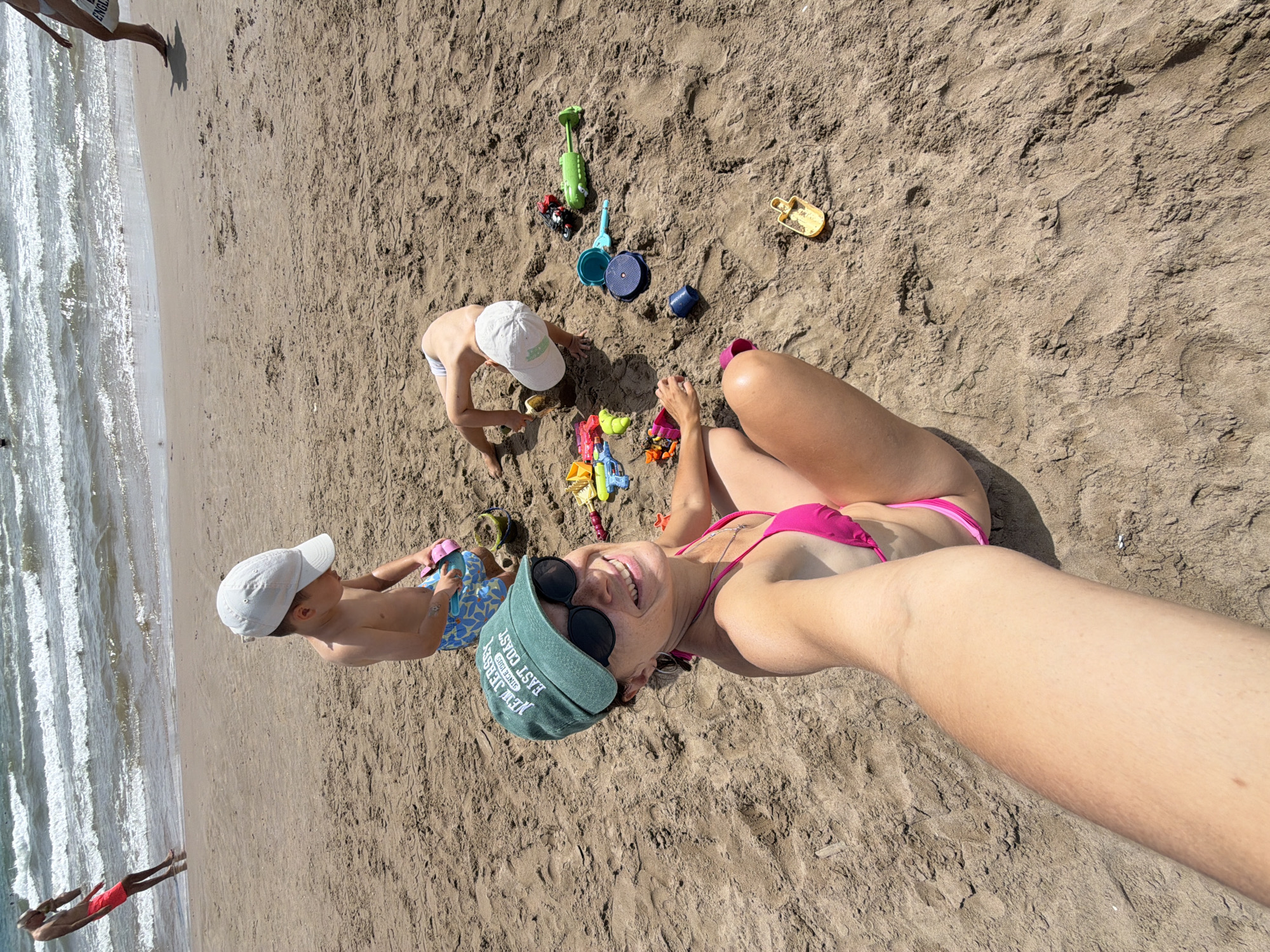 A woman in a pink bikini and a green baseball cap is taking a selfie on a sandy beach. Her left arm extends towards the camera, partially obscuring the foreground. She is smiling broadly and wearing sunglasses. Behind her, two young children are playing in the sand. One child, wearing a white cap and blue patterned swim trunks, is kneeling and looking down at a pink toy. The other child, wearing a white cap, is on their hands and knees, playing with sand toys. Scattered around the children are various colorful plastic beach toys, including shovels, buckets, and a watering can. The ocean with moderate waves is visible in the background, with a person in red shorts walking near the water's edge. The overall scene depicts a casual day at the beach with family.