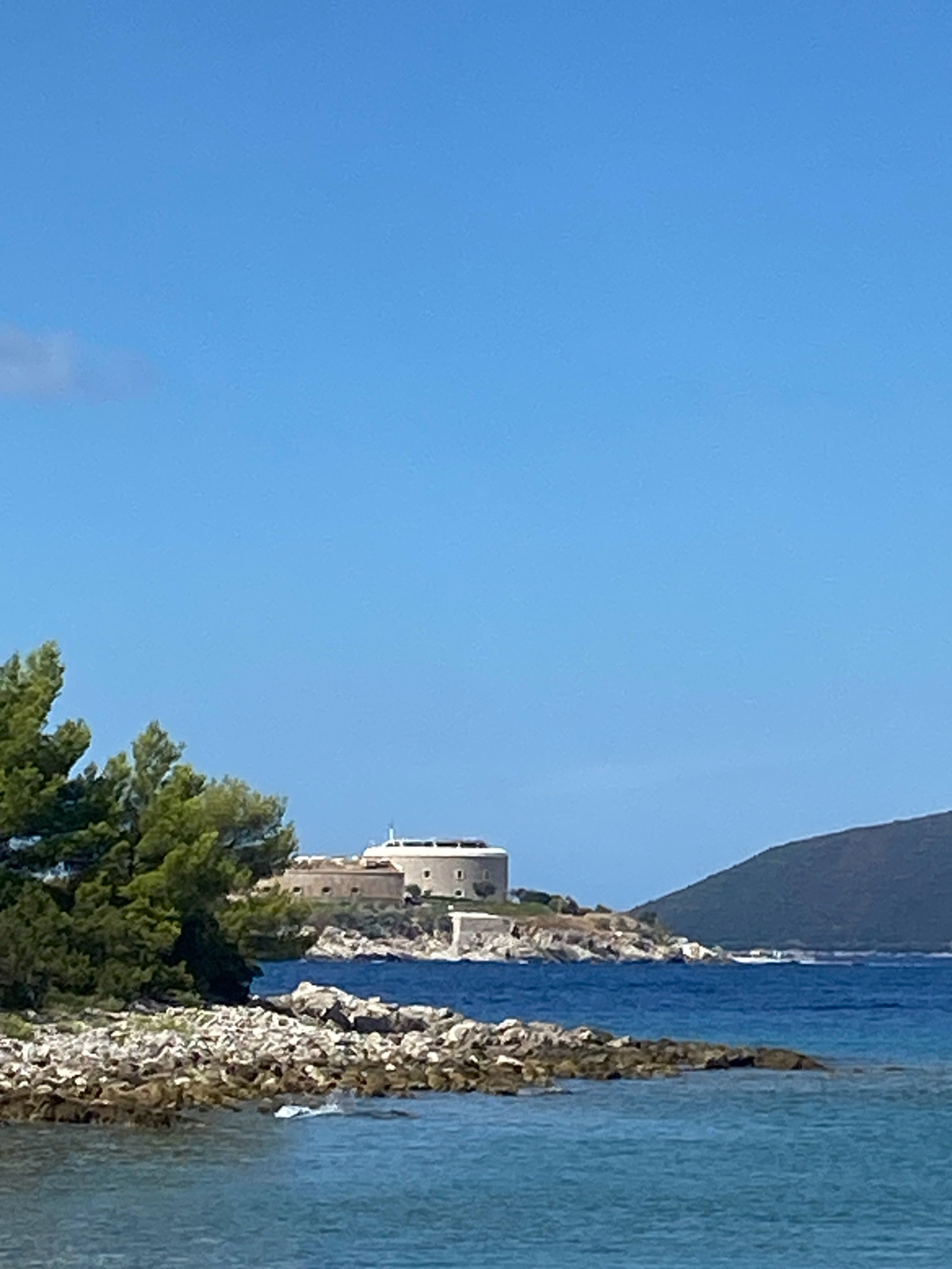 A circular fort with a white spire on top sits on a rocky outcrop of land jutting into a clear blue sea. The fort is surrounded by more rocky terrain and some greenery, including pine trees in the foreground. A dark, hilly landmass is visible in the background across the water. The sky is a bright, cloudless blue. The location is likely Forte Mare in Herceg Novi, on the Luštica peninsula of Montenegro.