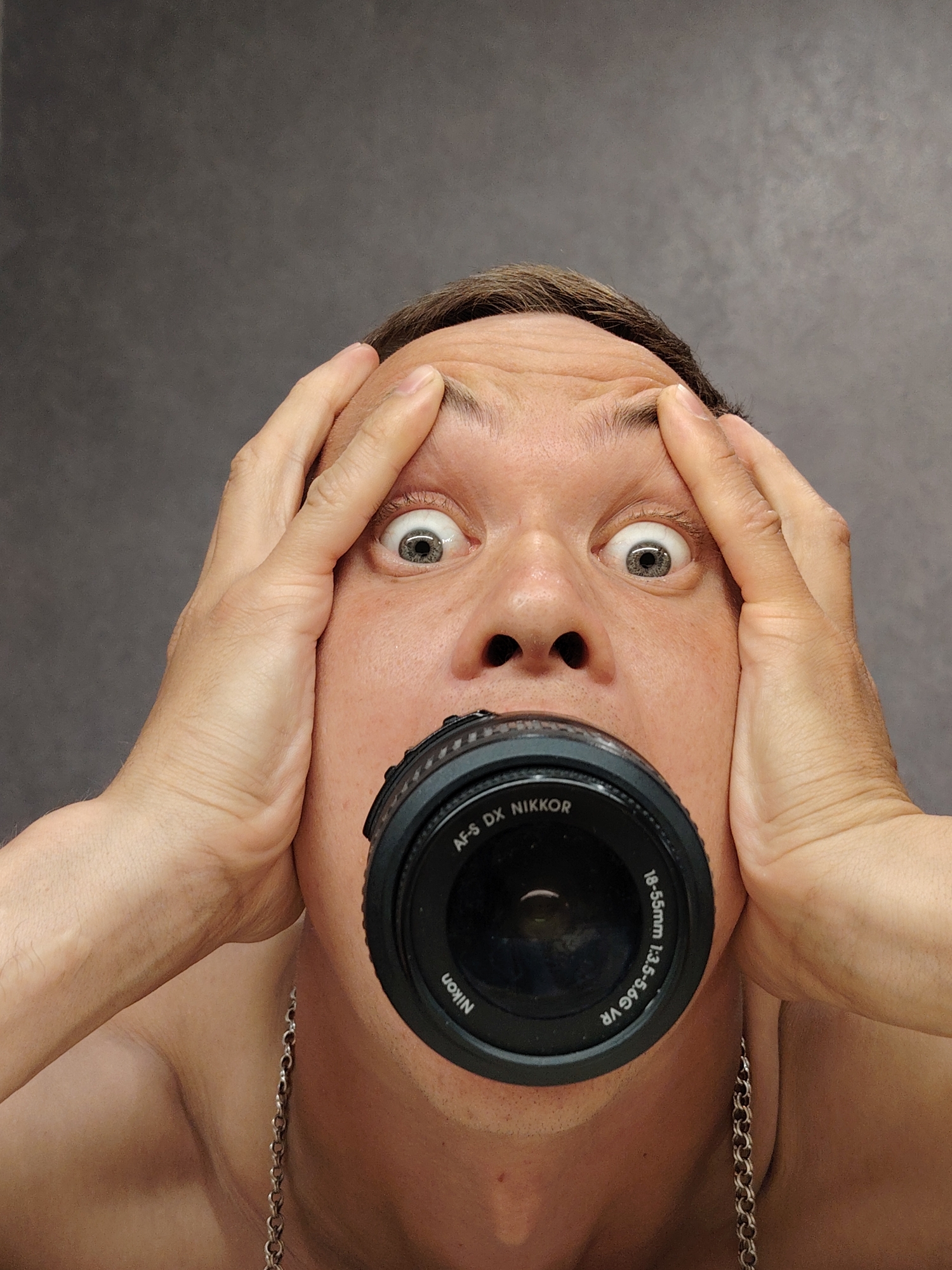 A close-up shot features a shirtless Caucasian man with brown hair and a chain necklace around his neck. He has a Nikon camera lens in his mouth, and his hands are pressed against his forehead with wide eyes, conveying an expression of shock or stress. The background is a plain gray wall. There is no indication from the image that the location is Chaikovsky, Russia.