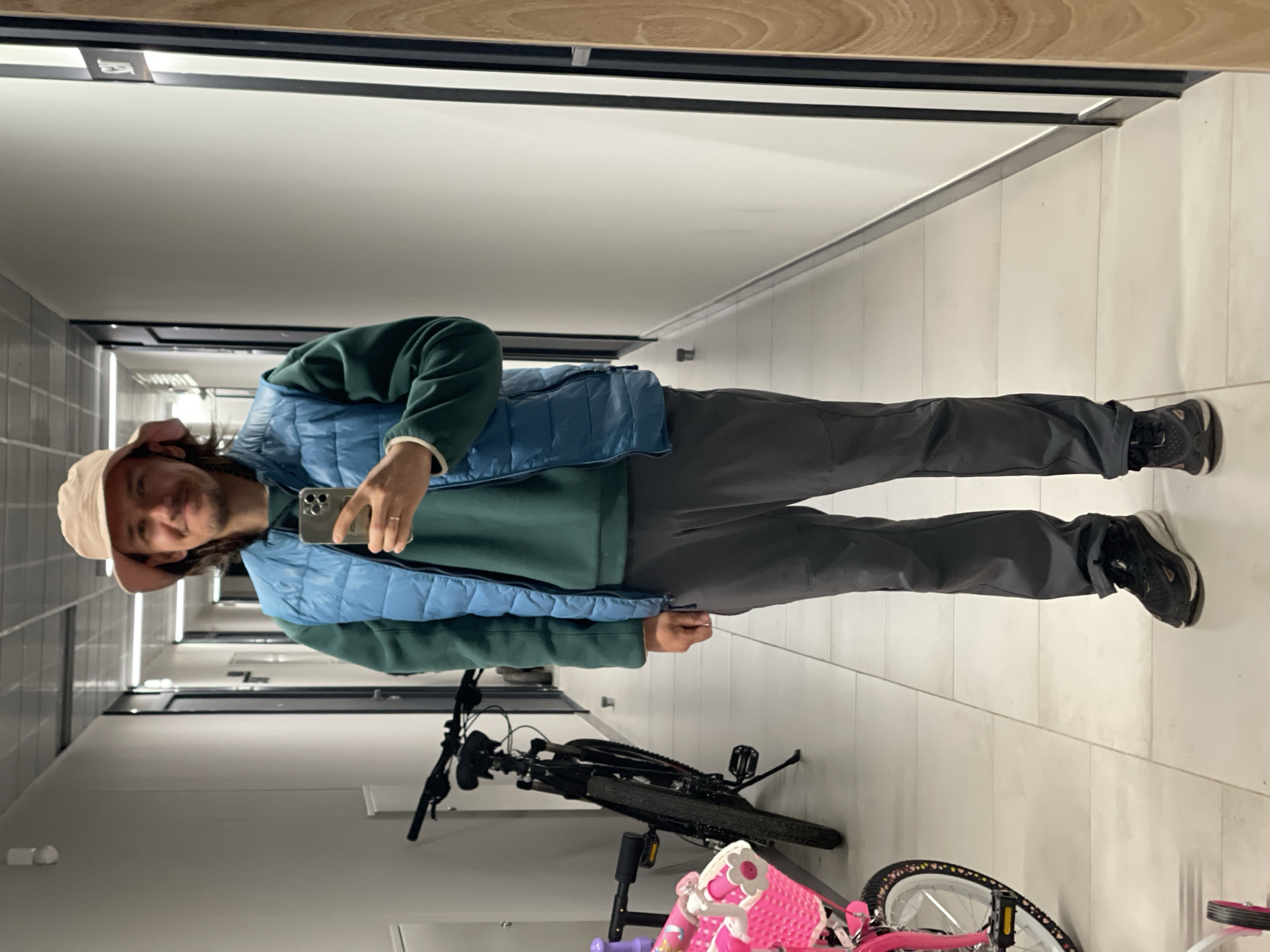 A man in a green sweatshirt, blue vest, gray pants, and a tan bucket hat stands in a hallway, holding a cell phone. He is looking at the phone's screen. There are two bicycles next to him. The location is a hallway in a city named Unknown, Unknown. The walls of the hallway are gray and white, and the floor is white tile. The lighting is fluorescent.