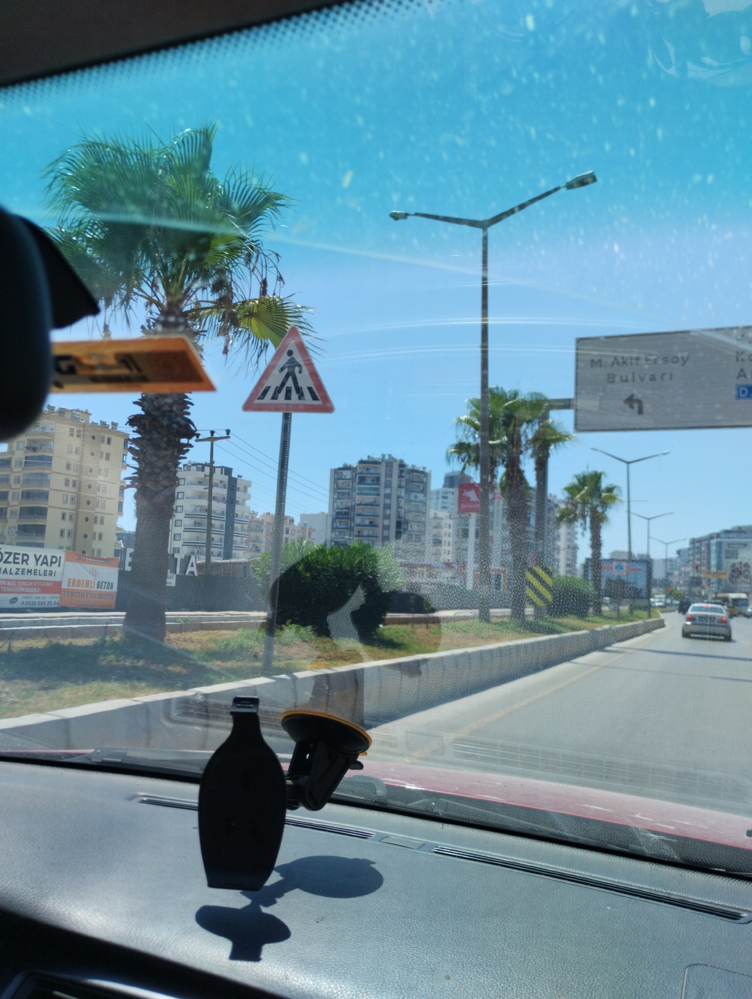 The image captures a view from inside a vehicle, looking through the windshield in Mezitli, Turkey. A black car mount is prominently displayed on the dashboard in the foreground. Outside, a street scene unfolds with palm trees and several multi-story buildings lining a road. A pedestrian crossing sign is visible, indicating a crosswalk. Traffic is light, with a few cars on the road. The sky is a clear blue, suggesting a sunny day. The overall environment appears to be an urban setting in Mezitli, characterized by modern architecture and Mediterranean vegetation.