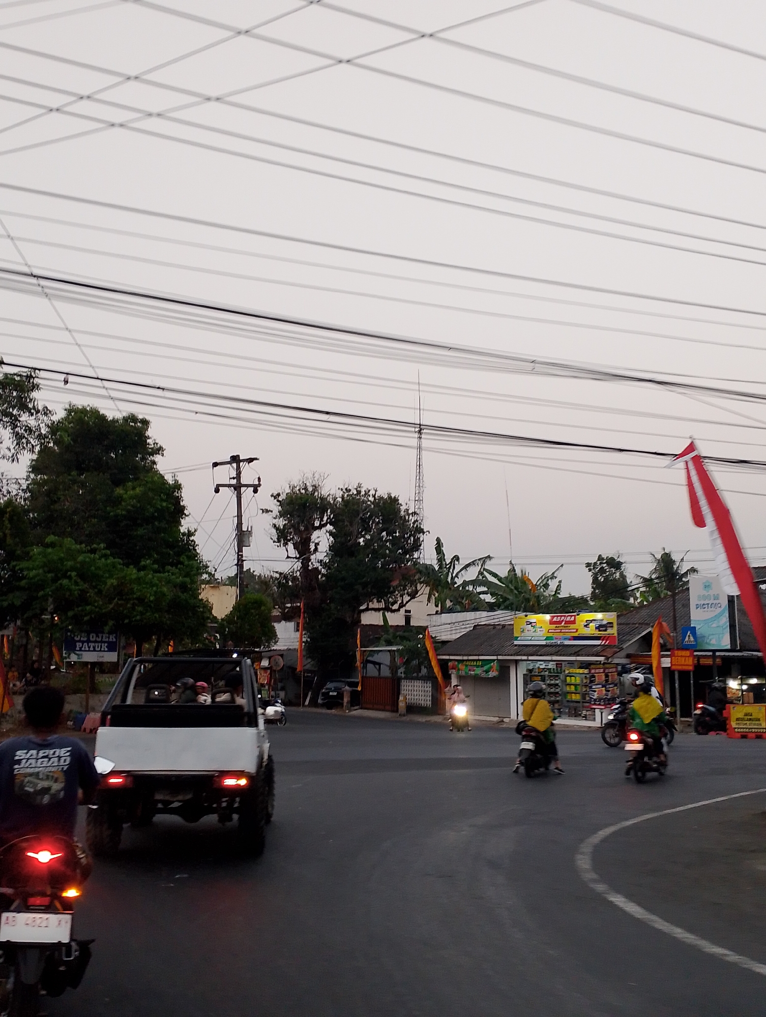 The image captures a street scene in the city of Patuk, Indonesia, likely during the late afternoon or early evening. The sky is overcast and appears a pale gray. Overhead, a dense network of power lines crisscrosses the frame, supported by utility poles. On the left side of the image, lush green trees provide a natural backdrop, with one prominent electrical pole visible among them. In the foreground, a white, open-top vehicle, possibly a Suzuki Jimny, is driving away from the camera. It has its rear brake lights illuminated, indicating it is actively moving. The vehicle's occupants are partially obscured. To the left of this vehicle, a person on a motorcycle, seen from behind, is also illuminated by their red tail light, and their license plate reads "AD 4921 XY".

The road surface is asphalt, with clear white lane markings visible. On the right side of the road, a row of shops is visible. One shop displays a prominent yellow and red advertisement for "ASPIRA BATTERY". Next to it, a blue sign with white lettering reads "200 M PICTO". Red and white Indonesian flags are also displayed, one prominently on the right side of the image, adding a patriotic element to the scene. Several other motorcycles are present on the road, with riders wearing helmets and casual clothing. Two riders on motorcycles are particularly noticeable in the mid-ground; one is wearing a yellow jacket and the other a green jacket. They appear to be navigating the intersection. The overall atmosphere suggests a typical urban street with ongoing traffic and commercial activity.