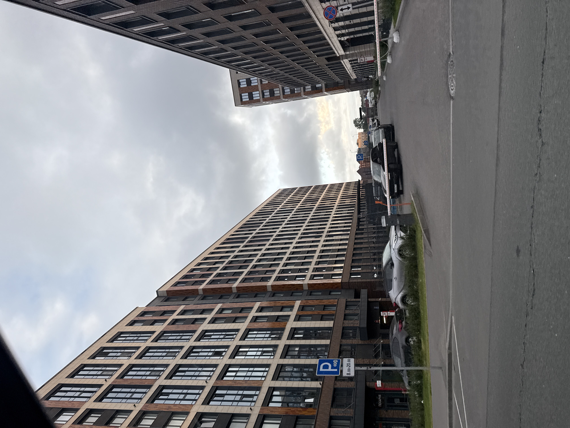 The image depicts a street scene in Saint Petersburg, Russia, characterized by modern, multi-story apartment buildings lining both sides of the road. The buildings are constructed with a combination of light-colored stone or composite panels and darker brick or metal accents, featuring numerous rectangular windows.
On the left, a light-colored building dominates the foreground, with a sign indicating "ПЕКАРНЯ" (Bakery) visible at its base. A blue parking sign with the letter "P" and additional text stating "8.00-20.00" is mounted on a pole in front of this building. Several cars are parked along the curb or in front of the buildings, including a silver SUV on the left and a dark-colored sedan further down the street.
On the right, a darker brick building runs parallel to the first, its facade also characterized by rows of windows. A white barrier gate, commonly found at building entrances or parking areas, is extended across the road. A red and white circular sign with a diagonal line and an arrow is visible on a pole near the barrier.
The sky is overcast with clouds, suggesting a cloudy or possibly rainy day. The road surface appears to be asphalt. The scene conveys a sense of urban development and residential living in a contemporary city setting.