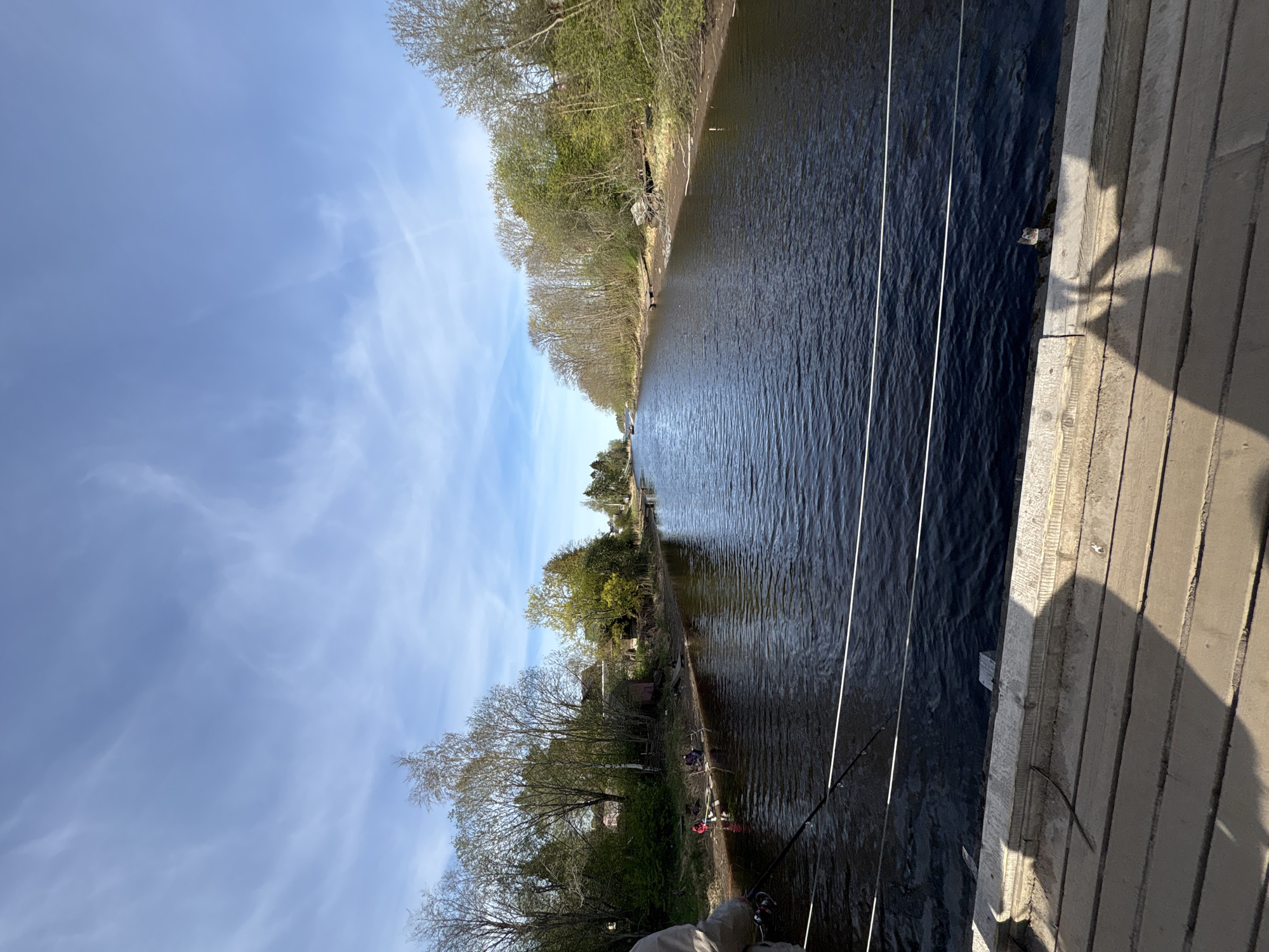 A view of a canal or river from a bridge, likely located in or near Приморский, Russia, given the query context. The water is dark and reflects the sky, and is flanked by treed banks on either side. On the right is a wooden structure, probably the bridge, with prominent shadows. Fishing lines are visible extending down into the water from the bridge. On the left side of the waterway, people are near the bank; they are presumably fishing or engaged in other waterside activities. The sky is light blue with wispy clouds.