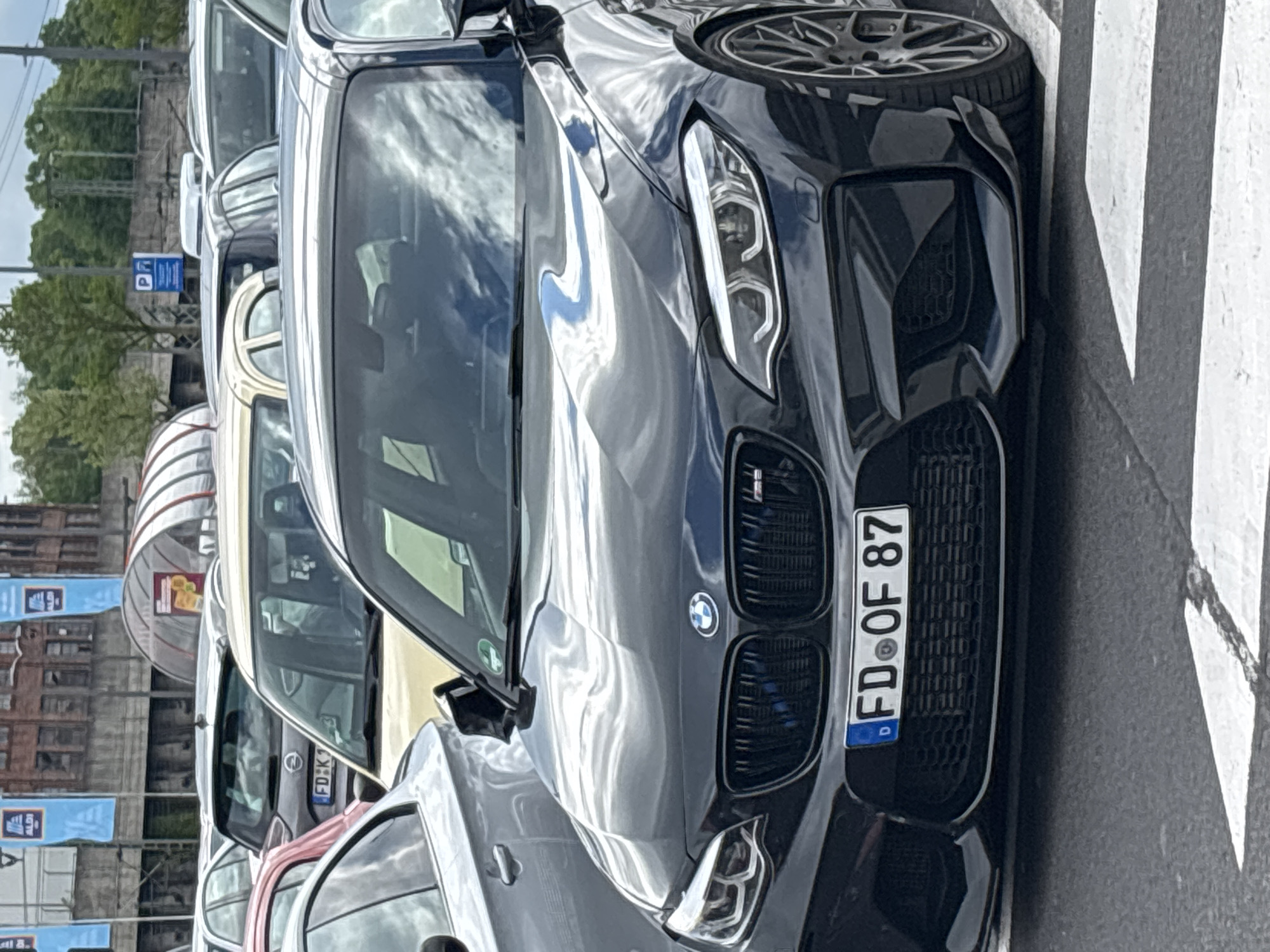 The image depicts a close-up of a dark-colored BMW parked on a street in Fulda, Germany. The car is in the foreground, with its front end and part of its driver's side visible. The license plate reads "FD OF 87," indicating it is registered in Fulda. The car is parked alongside other vehicles, including a light-colored Opel. In the background, banners with "ALDI" logos, buildings, and trees are visible. The sky is cloudy, and the reflections are visible on the car's hood and windshield. The car is parked on a crosswalk.