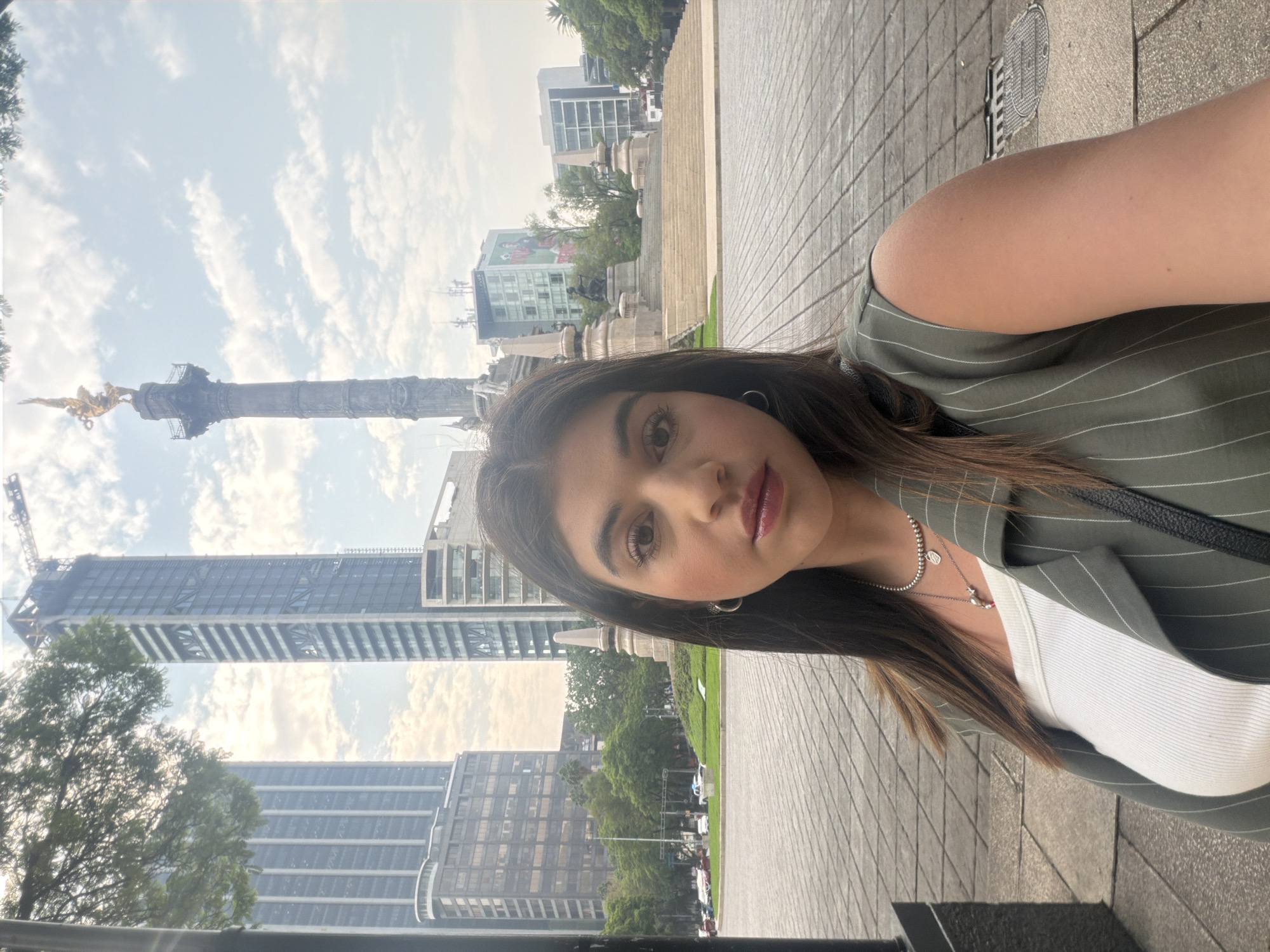 A woman is taking a selfie at the base of the Angel of Independence monument in Mexico City. The monument is a tall column topped with a golden statue of the angel, and skyscrapers and other buildings are visible in the background. The woman is wearing a green striped jacket and a white top, and she has dark hair and brown eyes. The environment includes a paved area, trees, and other urban elements.