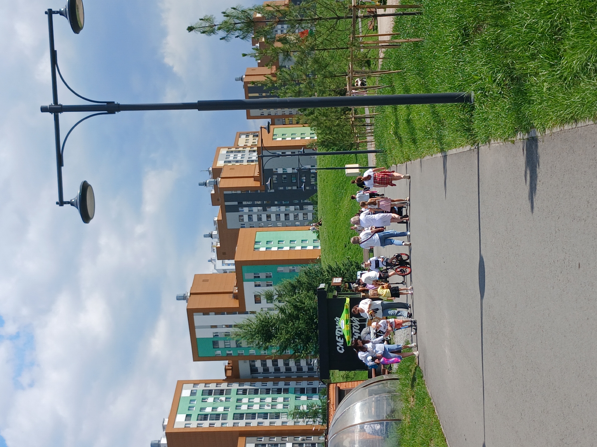 In Yekaterinburg, Russia, a group of people are walking along a paved pathway in a park-like setting with modern apartment buildings in the background. The buildings are characterized by their tall, multi-story structures with a mix of brick and green-colored facades. A prominent street lamp with two lights stands on the right side of the pathway.
On the pathway, a diverse group of individuals, including adults and children, are engaged in various activities. Several children are riding on scooters and a bicycle, while others walk alongside adults. One adult is pushing a stroller. A black kiosk with a green umbrella is visible on the left side of the pathway, displaying the word "СЛЕДУЮЩИЙ" which translates to "NEXT" in Russian. A transparent dome structure, possibly a greenhouse or observation area, is located to the left of the kiosk. The pathway is bordered by a lush green lawn and young trees with visible stakes for support. The sky is partly cloudy.
