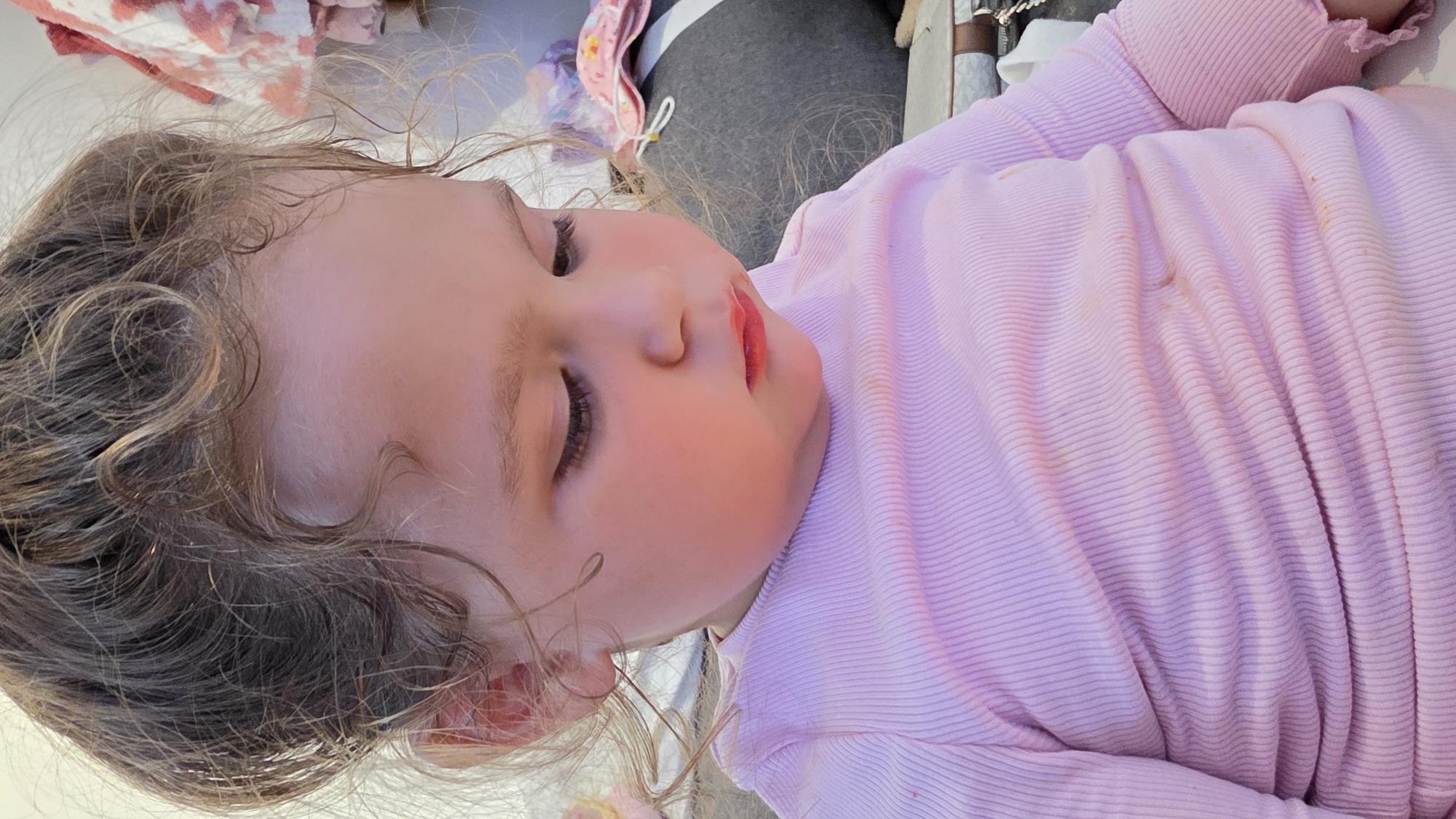 A young child with curly brown hair, fair skin, and closed eyes is lying down, wearing a pink long-sleeved shirt with stains. The background includes grey fabric, possibly clothing, and patterned fabrics in white, pink, and red. The image is taken from a close-up, slightly angled perspective. There is no indication of London, United Kingdom, or any specific location.