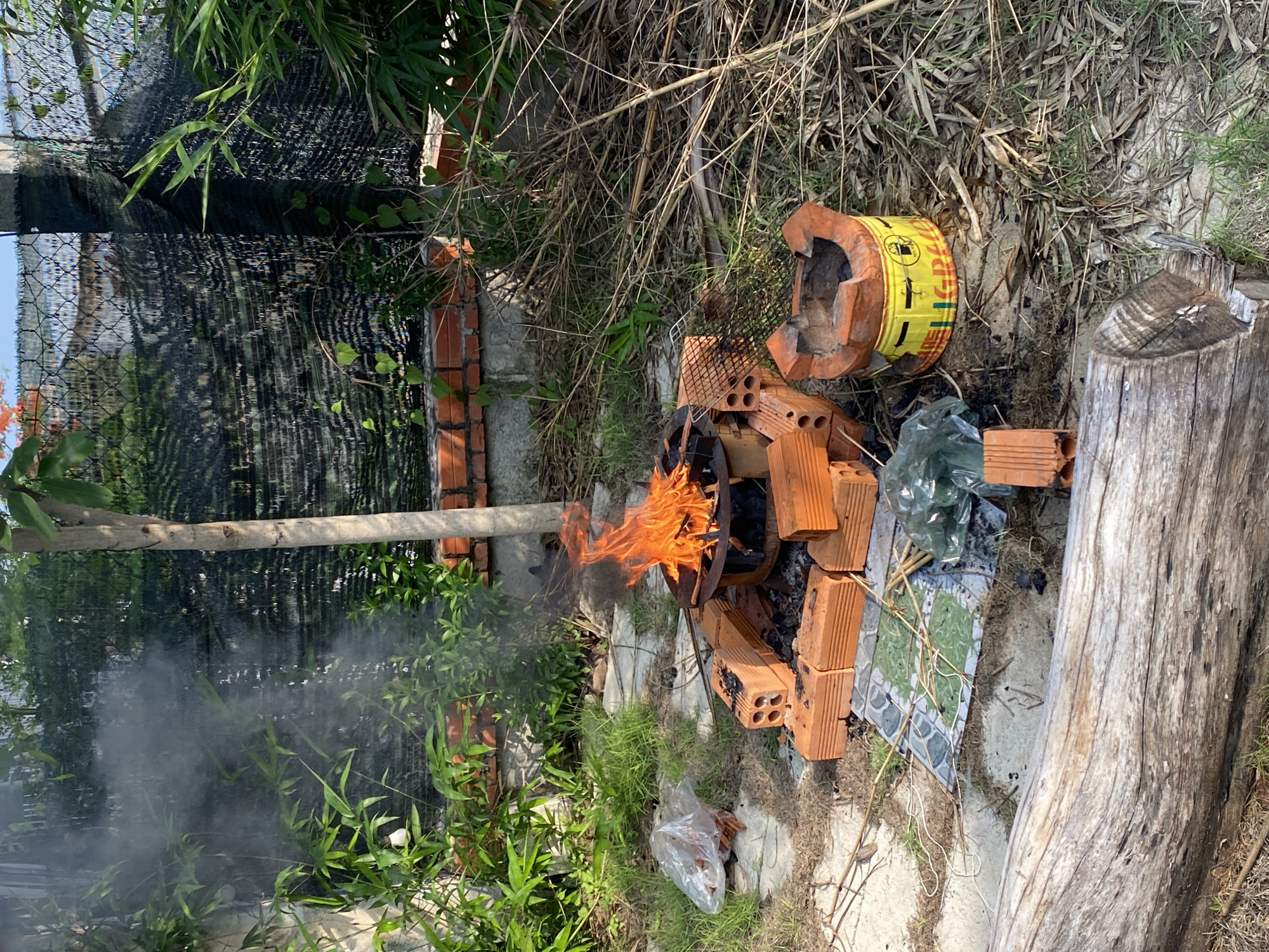 In Mũi Né, Vietnam, a fire burns in a makeshift outdoor cooking area. The fire is contained within a metal stove resting on a foundation of orange bricks. Surrounding the stove are more loose orange bricks, some showing signs of soot. To the left of the fire, a clear plastic bag contains what appears to be more bricks. In front of the fire, a patterned tile lies on the ground. Further to the right, a yellow cylindrical object with red and black writing, possibly another type of stove or container, sits on the sandy ground. The scene is set amidst dry grasses and vegetation, with lush green plants and a thin tree trunk to the left and rear. A black mesh fence is visible in the background, with some brickwork beneath it. A large, weathered log rests in the foreground. Smoke from the fire drifts upwards and to the left, mingling with the surrounding greenery and the fence.