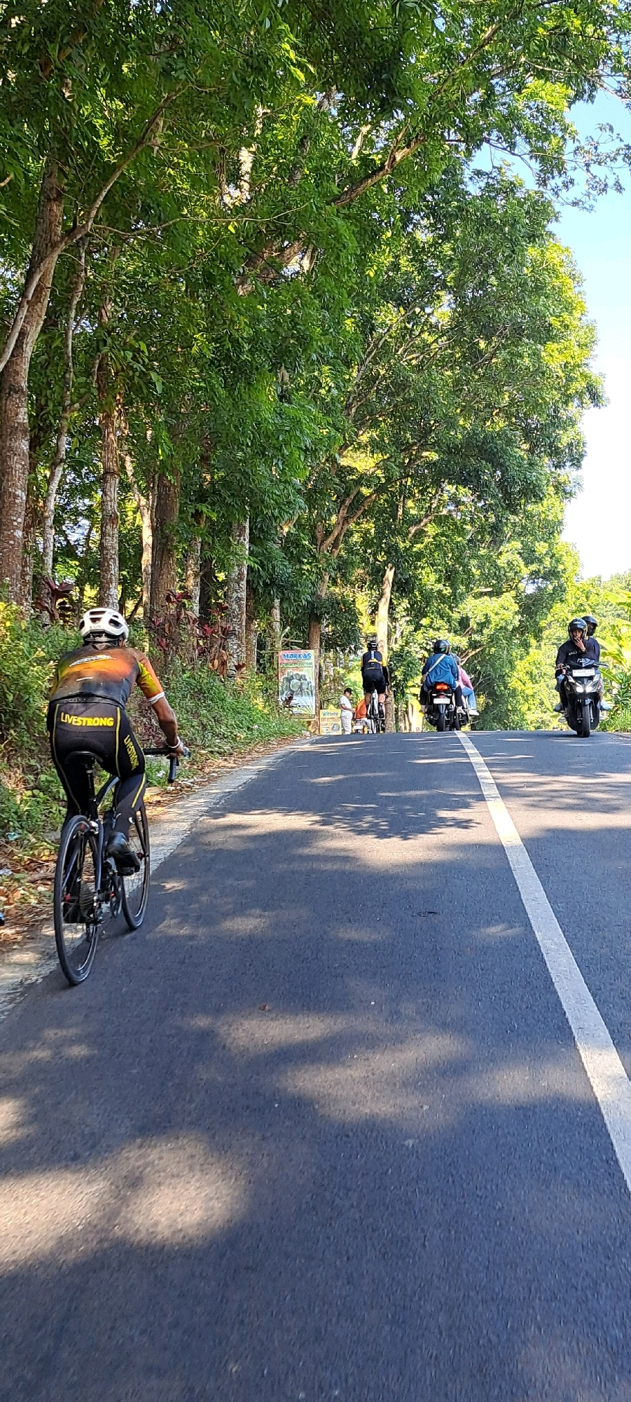 A cyclist wearing a black and orange jersey with "LIVESTRONG" text on the back is riding on a paved road with a white line down the center. They are wearing a white helmet and black cycling shorts. The road is bordered by lush green trees on the left and right. In the distance, several motorcycles are also on the road. On the right side of the road, a black motorcycle with two riders is visible, and further ahead, another black motorcycle with a single rider. On the left side of the road, a sign with text and images is posted. The scene appears to be in a rural or semi-rural area, possibly in or near Belik, Indonesia, given the vegetation and the presence of motorcycles. The lighting suggests it is daytime, with sunlight casting shadows on the road from the trees.