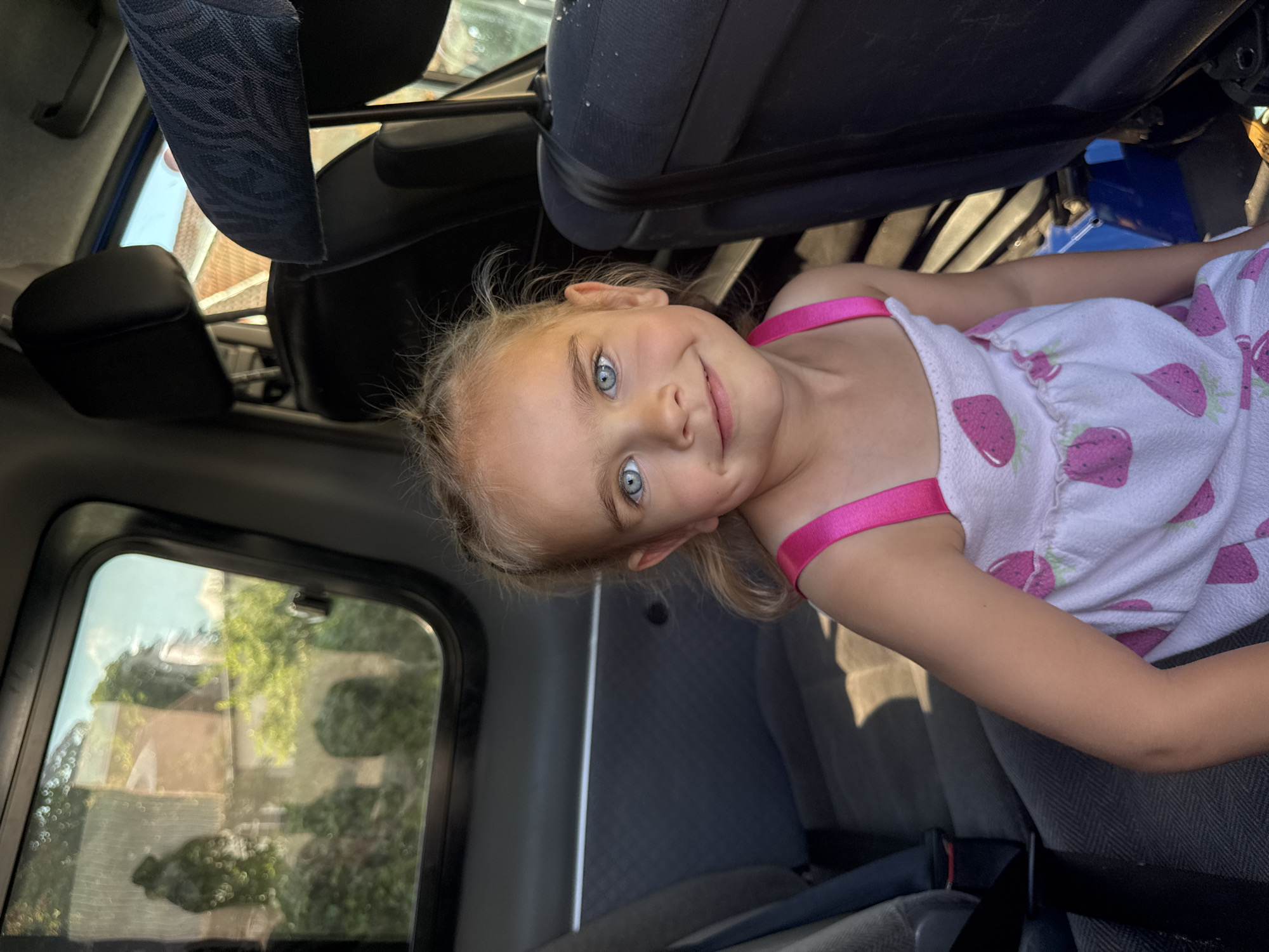 A young girl with bright blue eyes and blonde hair is lying down inside a vehicle, possibly a car or van. She is wearing a white dress with pink strawberry patterns and pink straps. Her head is tilted, and she is looking at the camera with a cheerful expression, a slight smile on her face. The interior of the vehicle is visible, with dark-colored seats and a window on the left side showing a blurred view of trees and buildings, suggesting an outdoor, daytime setting. The lighting indicates it is daytime. The girl appears to be relaxed and happy. There is no visible text in the image.