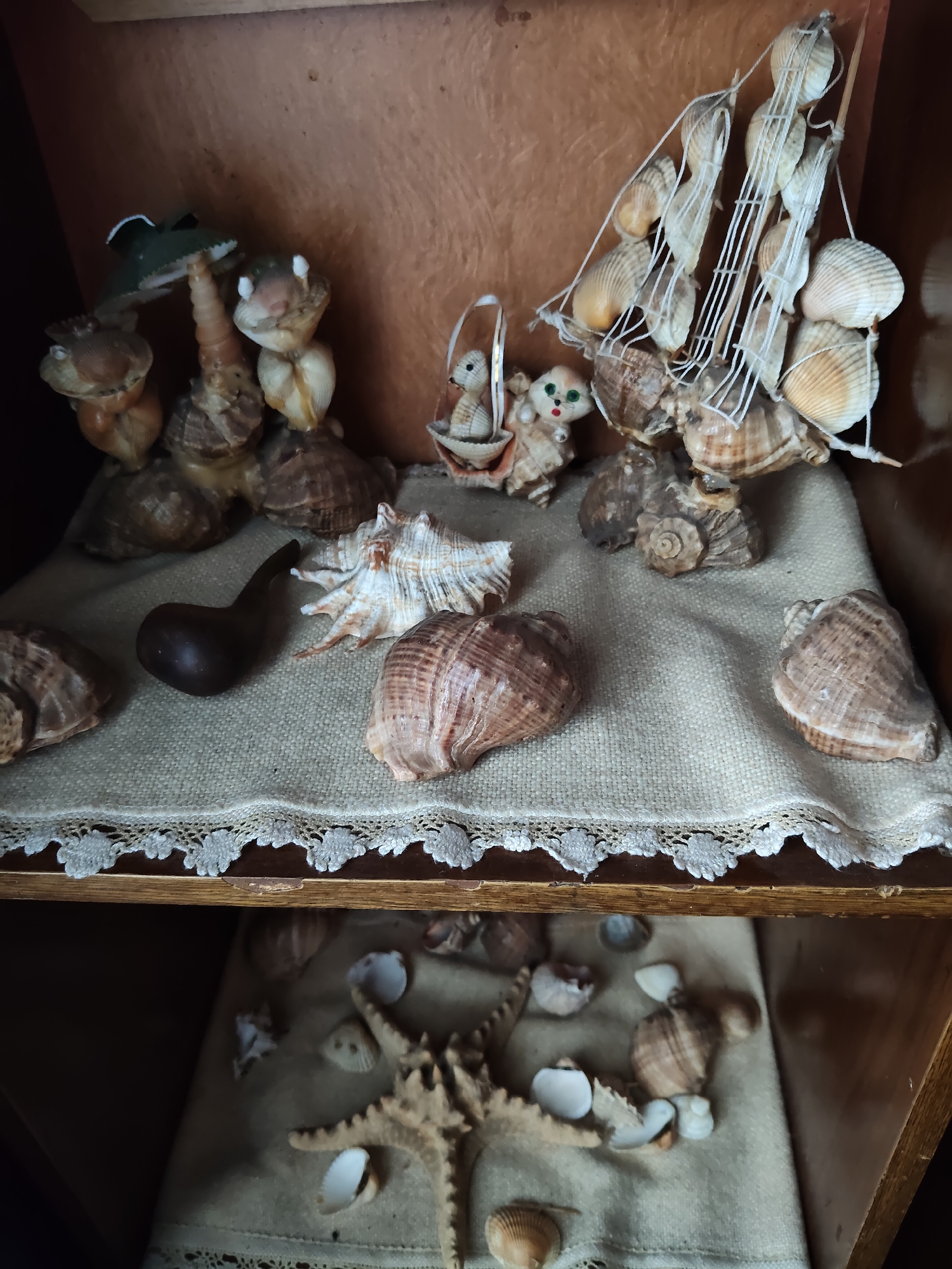 The image shows a shelf in a dimly lit room, likely a home interior in Gîrbova, Moldova. The shelf is decorated with various seashells arranged to form decorative items. Prominently displayed is a model sailboat made of shells and twine, situated behind a group of shell figures resembling animals, including a frog and two small creatures with ears and eyes. In front of these figures, a dark, curved object, possibly a pipe, lies on a beige fabric with a lace trim. Several other large seashells are scattered on the fabric, some appearing to be conch shells. The shelf itself appears to be made of wood. Below this shelf, another shelf is visible, also covered with fabric and displaying a starfish and numerous smaller seashells. The overall impression is one of a personal collection of maritime-themed decor.