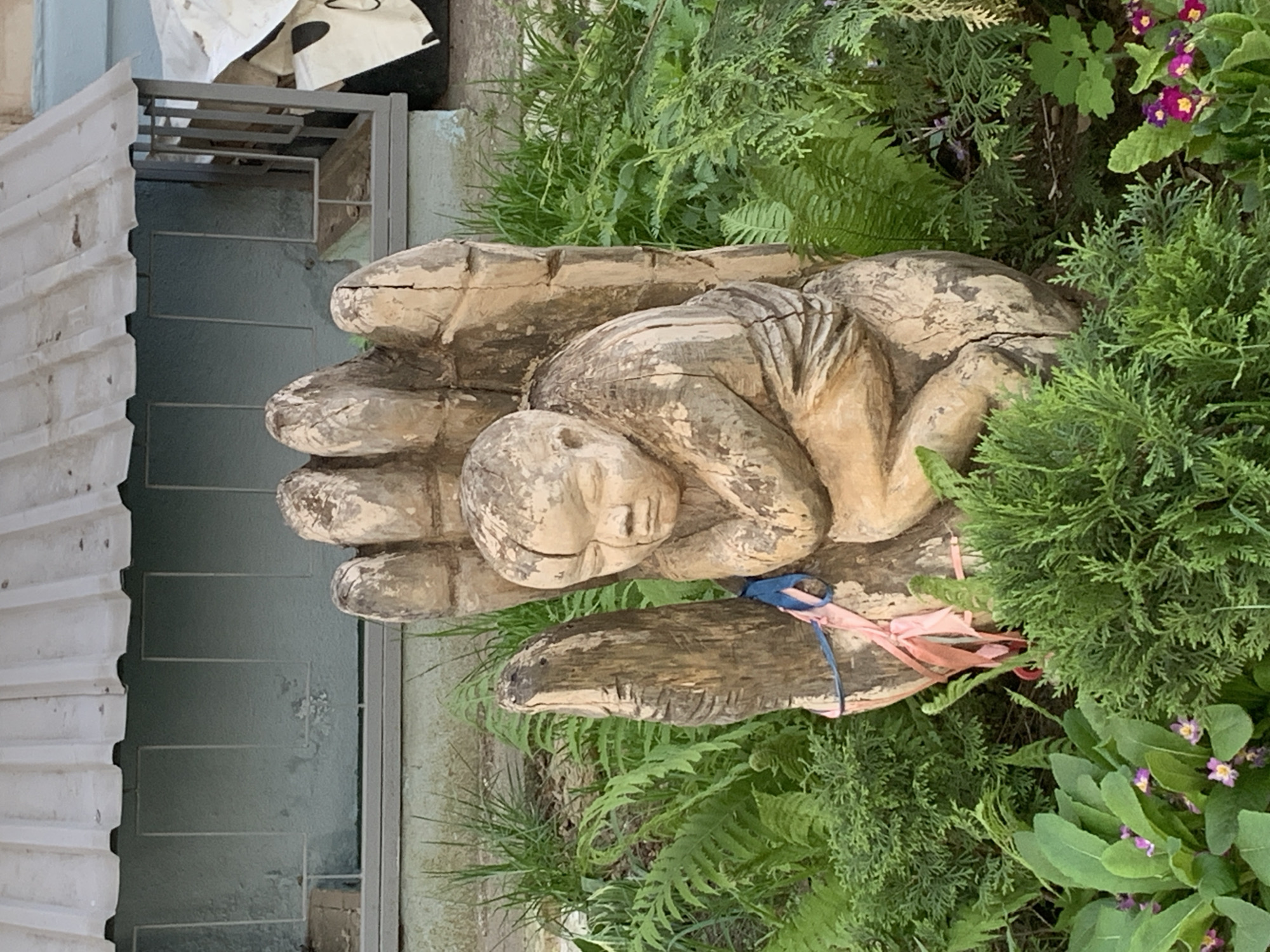 A carved wooden sculpture of a person curled up in a hand is nestled among ferns, bushes, and flowers. A light blue and pink ribbon is tied around the wrist of the wooden hand. The sculpture is located outdoors next to a building with a grey door and a metal gate. The sculpture's location is in the city of Chaikovsky, Russia.
