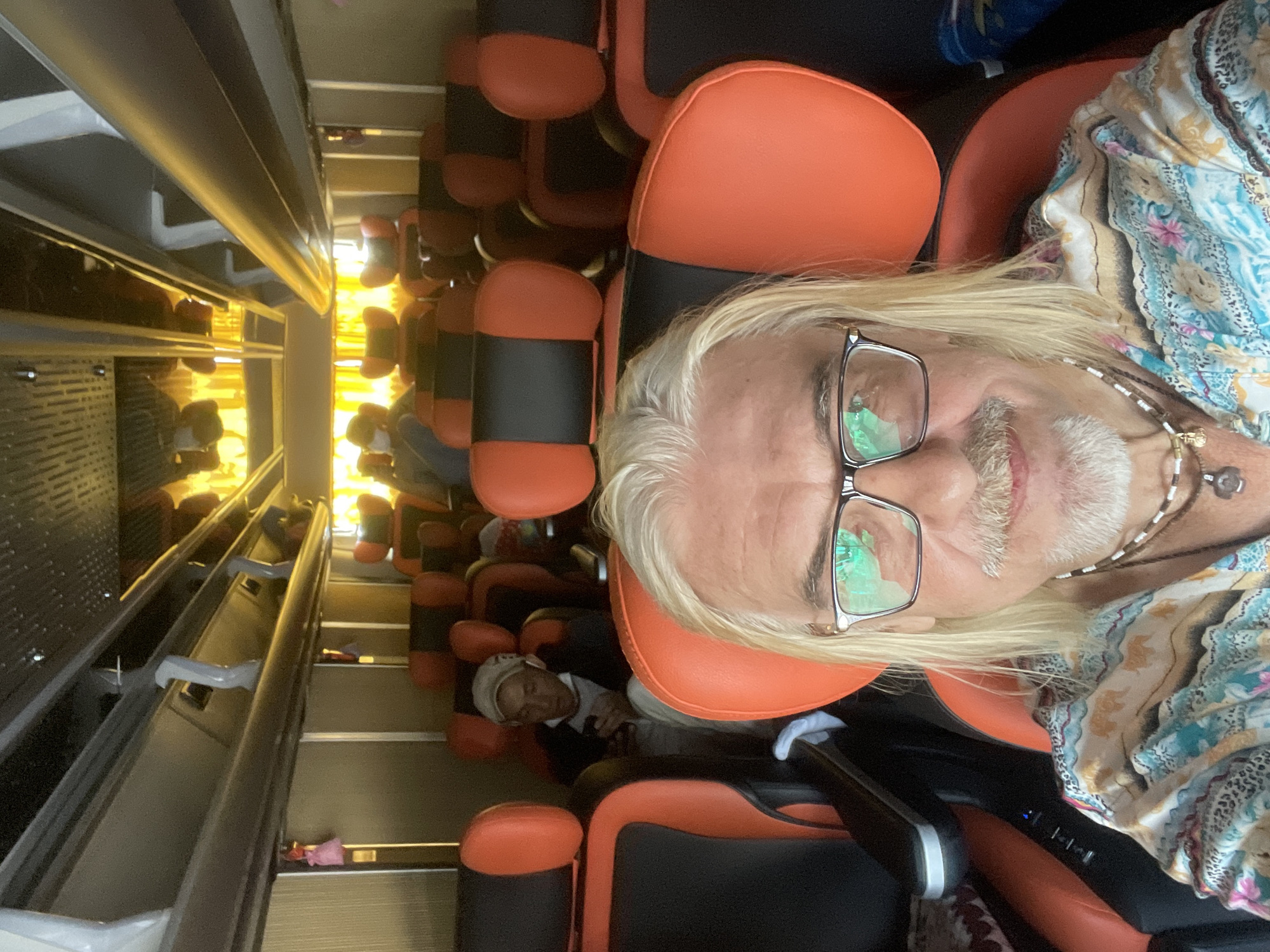 The image shows a man with long blond hair and a mustache taking a selfie inside a bus. He is wearing glasses, a colorful patterned shirt, and two necklaces. The bus has orange and black seats. In the background, other passengers can be seen sitting in their seats, and some are using their phones. The bus appears to be in motion, with yellow curtains visible through the windows. The man is in the foreground, with the bus interior extending behind him. The image does not provide specific details about the location in Phu Thuy Ward, Vietnam.