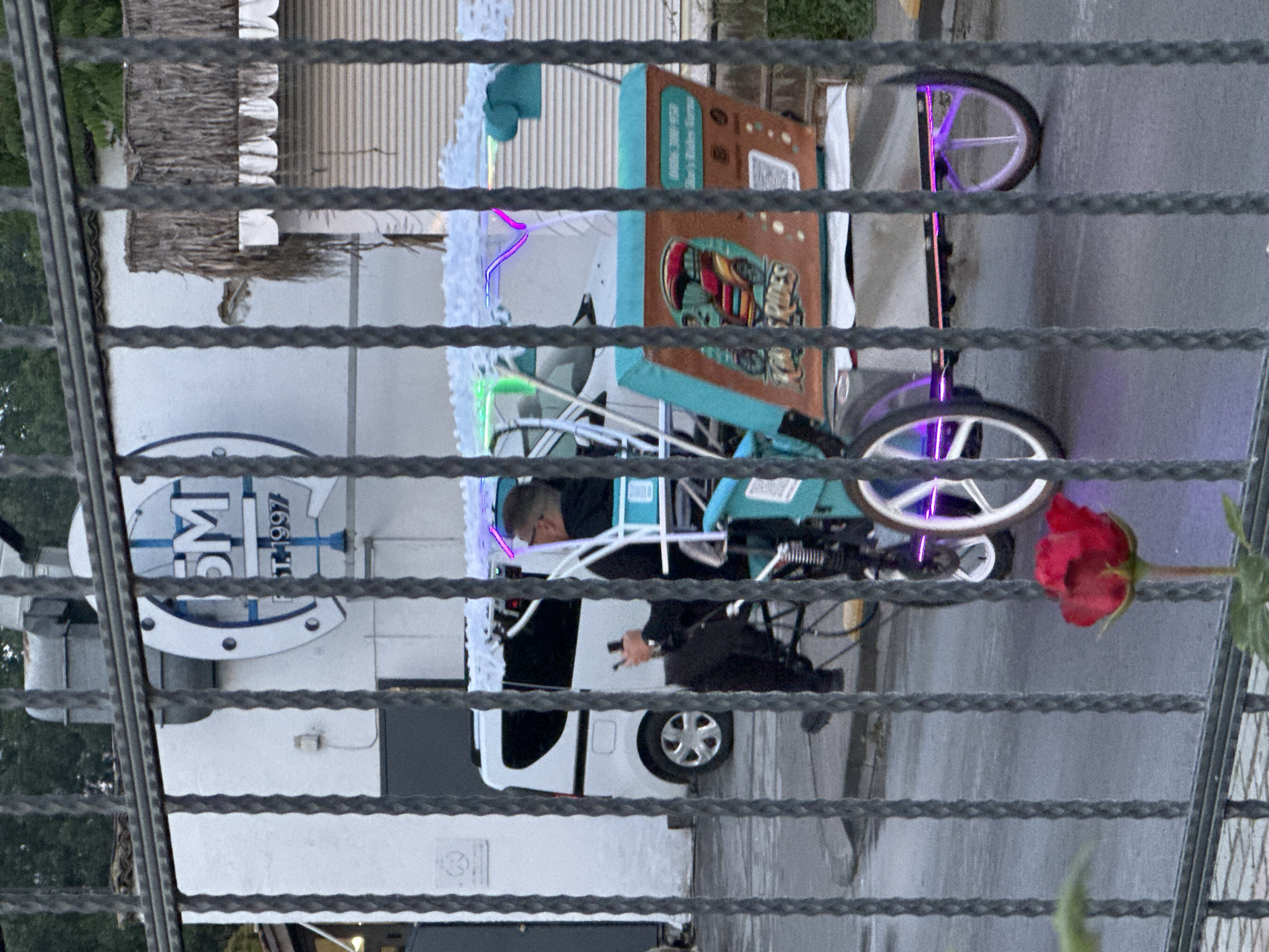 The image shows a street scene in Varna, Bulgaria, viewed through a metal fence. A man in a black outfit operates a decorated tricycle with illuminated wheels and neon lights. The tricycle has a cargo box on the back, displaying text and an image of "ESQUITES." Behind the tricycle, there's a white vehicle with a visible license plate. A business sign with "DM EST 1997" is attached to a building on the left. A red flower hangs from the right side of the fence.