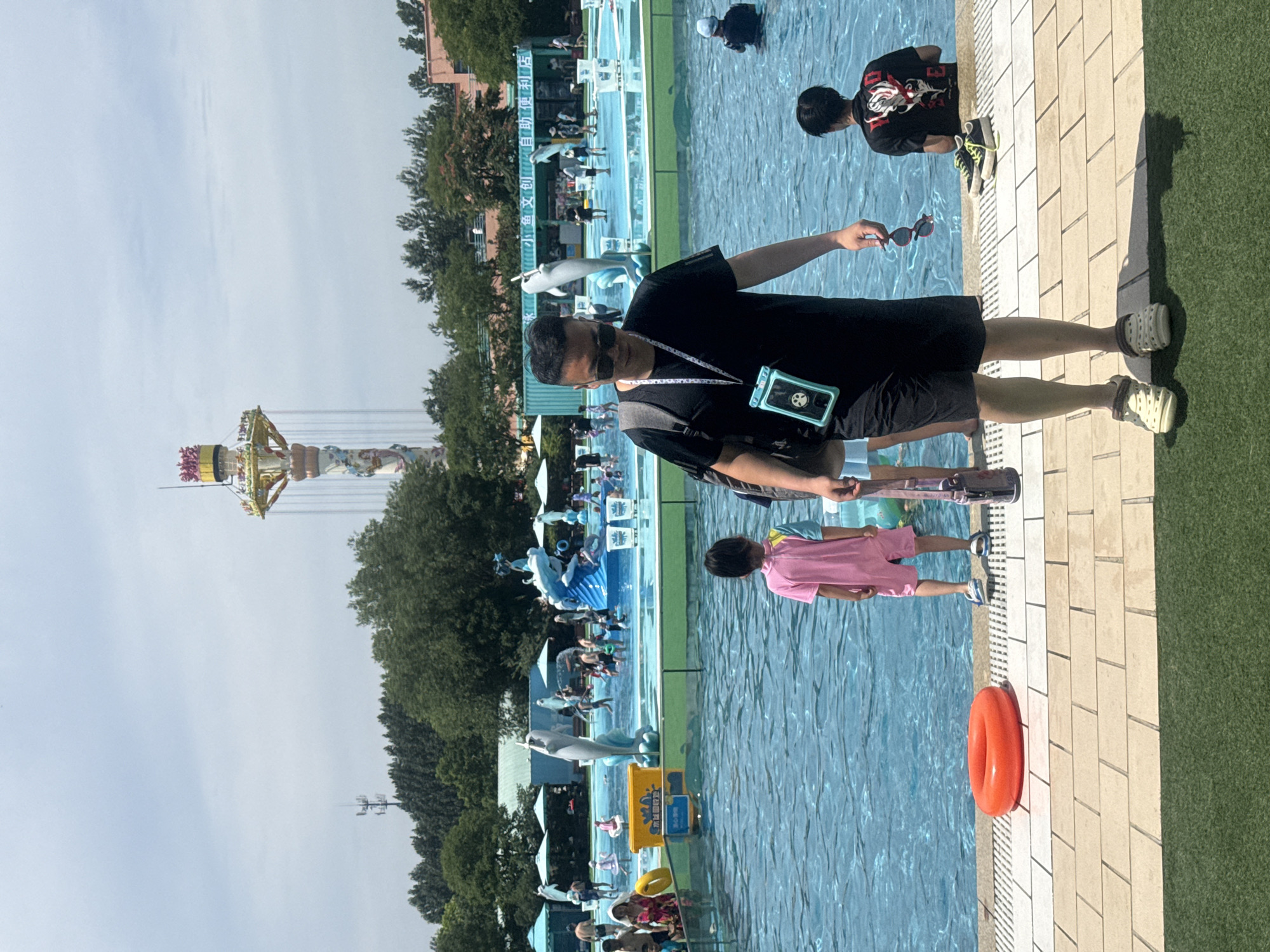 In Qinhuangdao, China, a water park is depicted on a sunny day. In the foreground, a man walks towards the camera, wearing sunglasses, a black t-shirt, black shorts, and light-colored clogs. He carries a dark backpack and a clear waterproof phone case around his neck. To his right, a young child in a pink and blue swimsuit stands facing away from the camera. Behind them, the pool is filled with people, including children and adults. Some are swimming, while others are standing in the shallow water. In the background, there's a large water slide with dolphin-themed decorations, as well as a tall amusement park ride. Trees and buildings surround the water park. An orange inflatable ring is visible on the tiled edge of the pool.