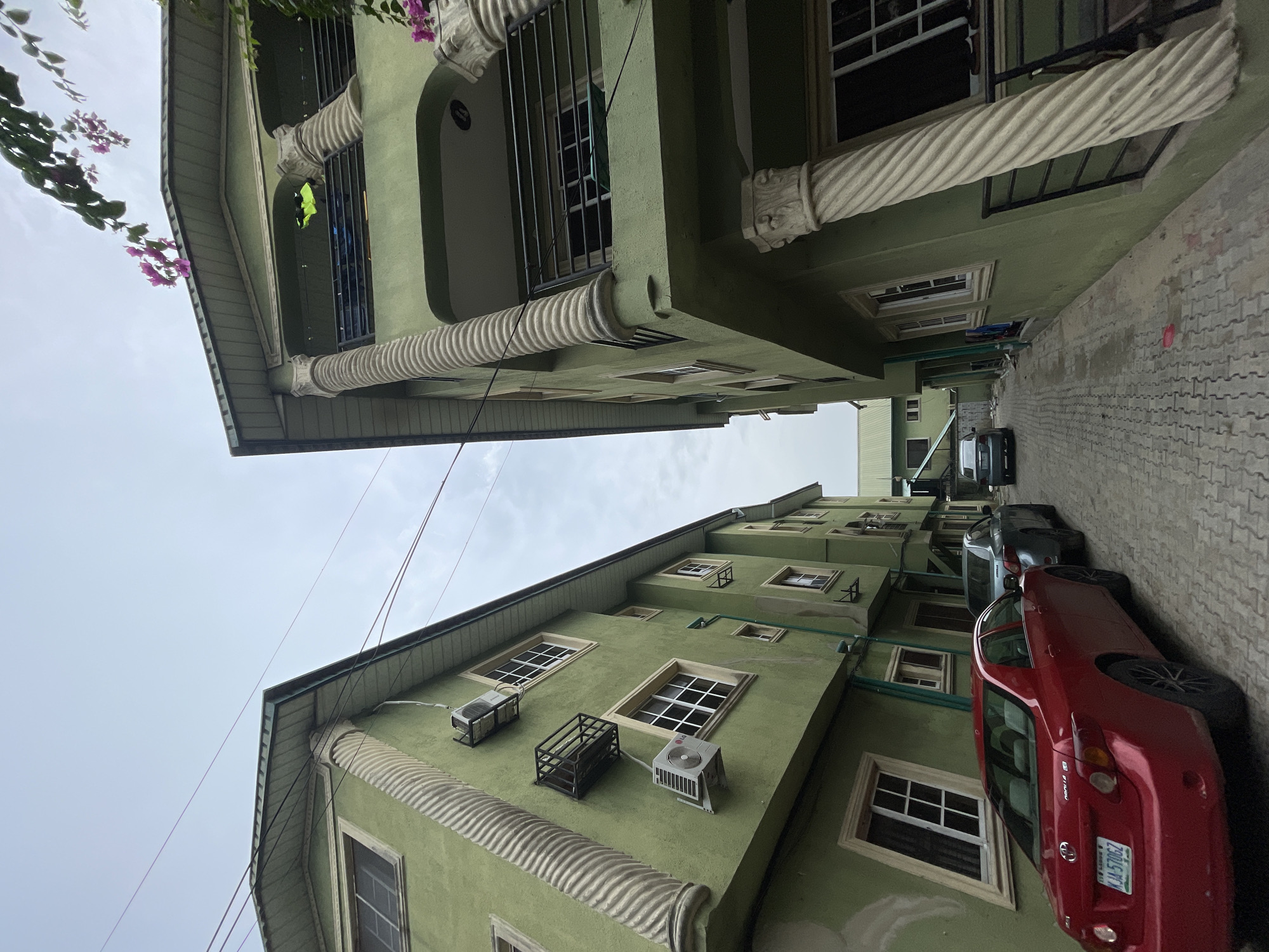 An eye-level shot taken in Ado, Nigeria shows two green buildings facing each other over a narrow street. Several cars are parked along the side of the building on the right, and AC units are mounted on the side of the building. The buildings have white, carved columns running along the sides, between window levels. Several windows, mostly covered with dark screens, are visible on both buildings. Wires run across the sky between the buildings.