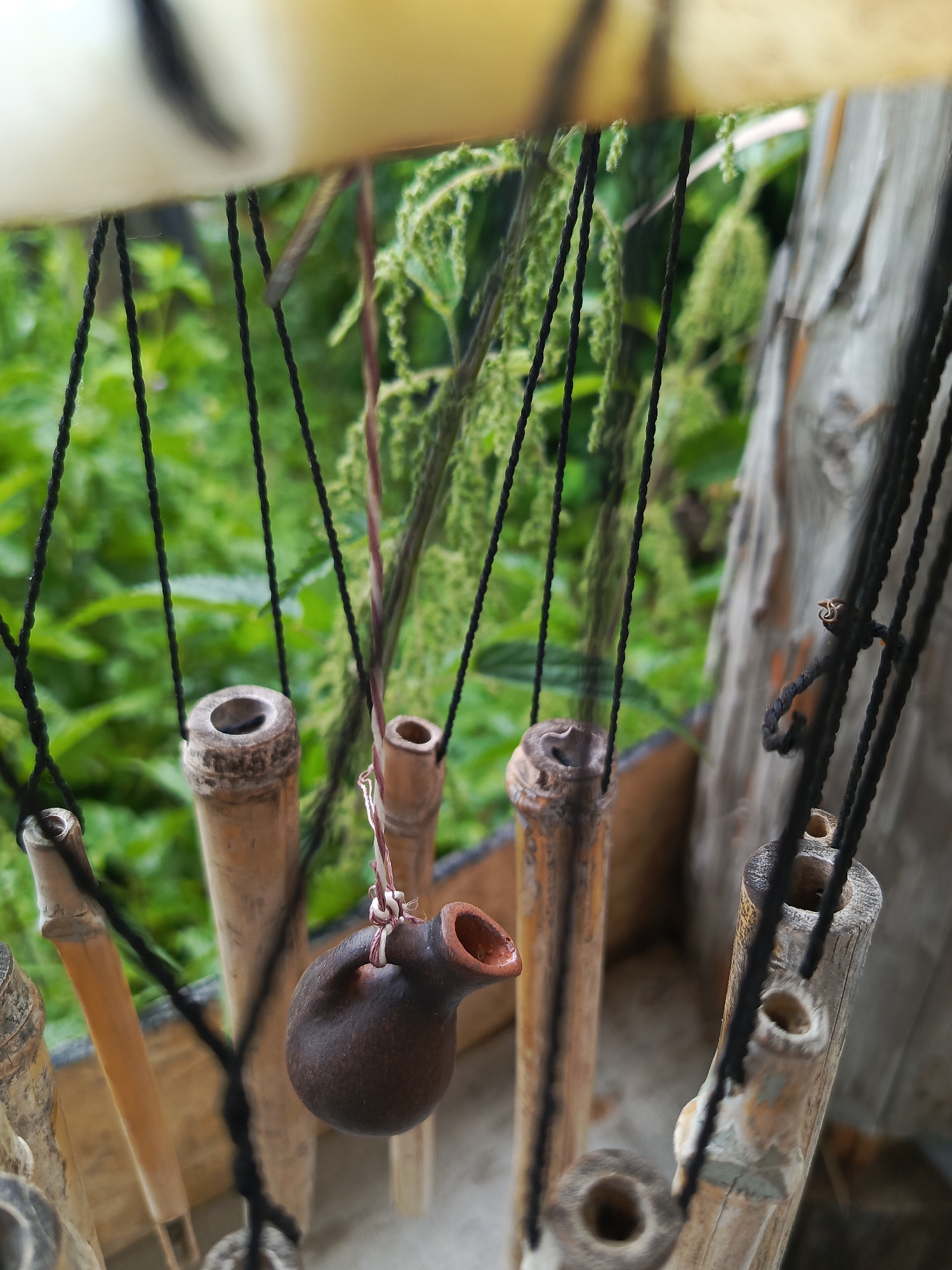 A close-up, eye-level shot showcases a handmade wind chime composed of bamboo tubes and a small, dark brown clay jug. The bamboo tubes, varying in thickness and length, hang vertically, suspended by black strings. The jug, with a narrow spout, is also suspended by a string and is positioned in the foreground, slightly to the left of the center. Its texture appears rough and unglazed. In the background, out-of-focus green foliage suggests an outdoor environment, possibly a garden or natural landscape. The light appears soft and diffused, indicating it might be a cloudy day or that the subjects are in a shaded area. The overall scene evokes a rustic, handmade aesthetic. The location is Kauchuk Stantsiya, Russia.