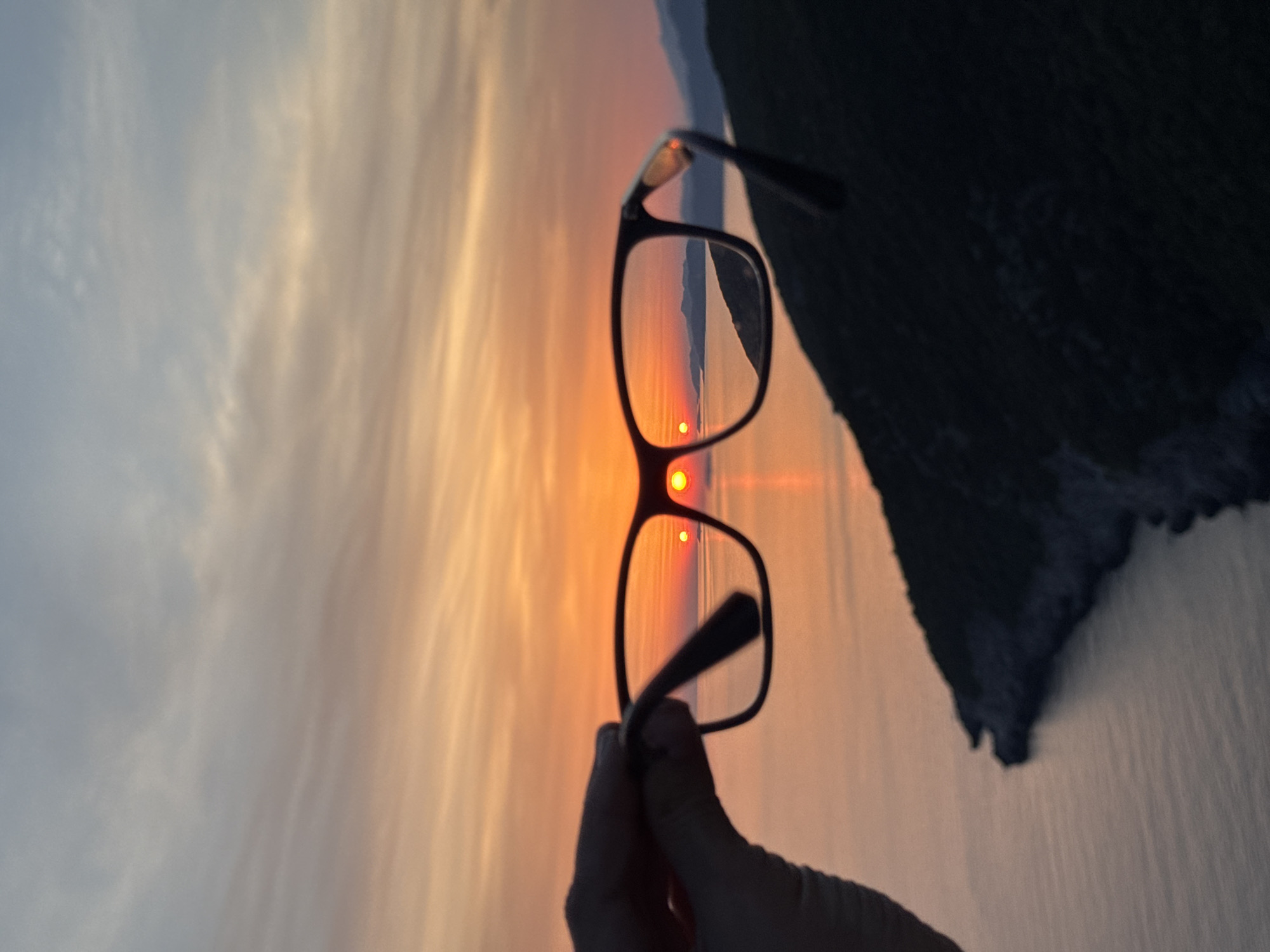 A pair of dark-rimmed eyeglasses is held in silhouette by a hand in the foreground. Through the lenses of the glasses, a vibrant sunset is visible, painting the sky in hues of orange, yellow, and pink. The sun appears as a bright orb, with smaller reflections or refractions visible within the lenses. Beyond the sunset, the calm sea stretches towards distant, silhouetted islands or mountains. The lower portion of the image features a dark, shadowy hillside, likely overlooking the sea. The scene suggests a coastal viewpoint, possibly in the region of Zagora, Montenegro, known for its scenic sunsets over the Adriatic Sea.