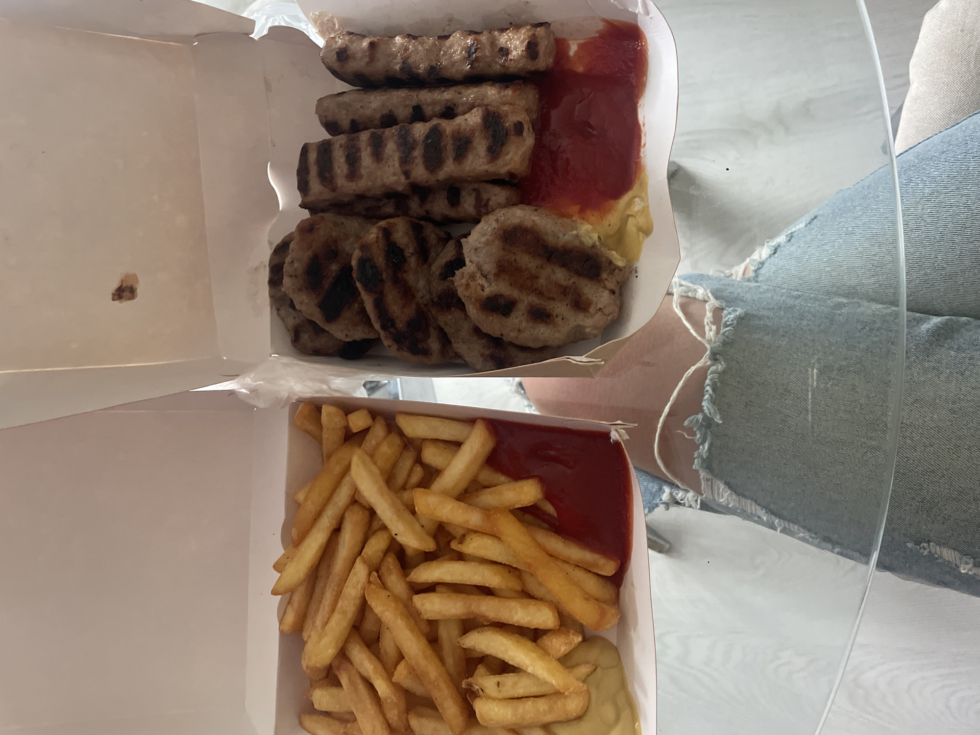 The image captures a top-down view of two open white cardboard boxes filled with a meal, likely takeaway, placed on a light-colored surface. A transparent surface, possibly a glass table, is prominent in the foreground and center, reflecting light.
The top box contains a generous portion of grilled meat. Several elongated, ridged pieces resembling ćevapi are stacked, alongside several flatter, round grilled patties, similar to pljeskavica or grilled burgers. On the right side of the box, there are dollops of two sauces: a vibrant red (ketchup) and a pale yellow (mustard).
The bottom box holds a large serving of golden-brown french fries. Similar to the top box, it also features dollops of red ketchup and yellow mustard on the right side.
Part of a person's body is visible on the right side of the frame, specifically a leg or thigh, clad in light-wash denim jeans with prominent rips and distressed areas, indicating a casual setting. The overall scene suggests an informal meal, possibly consumed at home or a relaxed environment. The type of grilled meat and fries with sauces is a common fast-food or casual meal option found in the Balkan region, including Sofia, Bulgaria. No text is visible in the image.