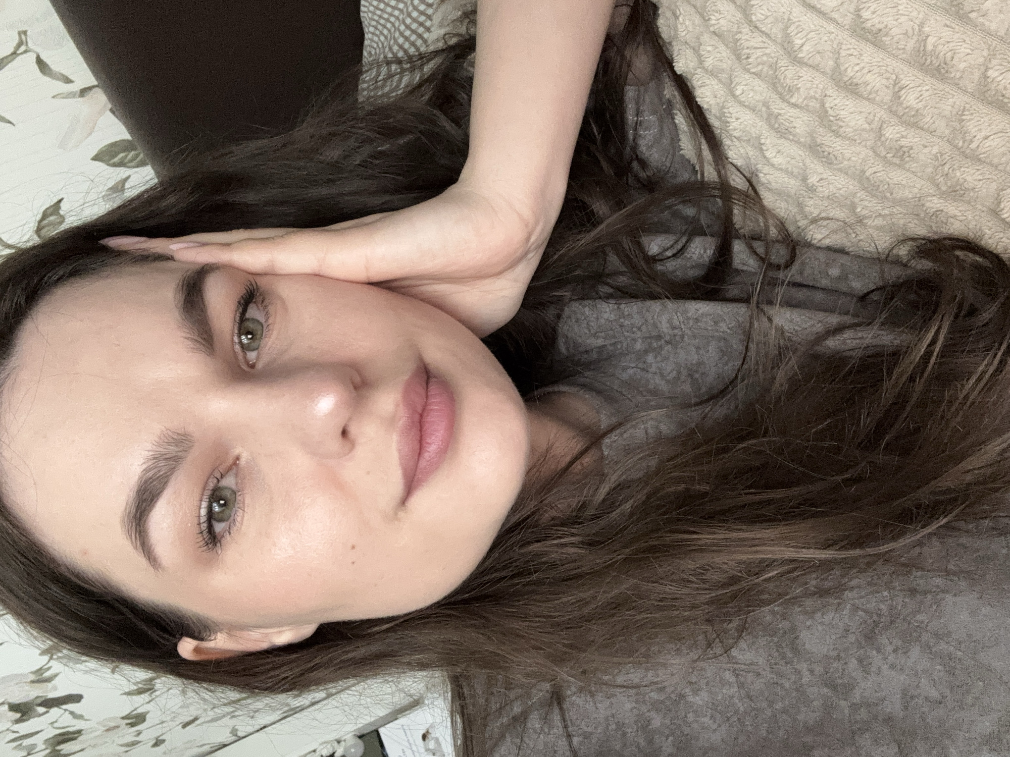 A woman with long, dark brown hair is lying on a patterned blanket, her head resting on her hand. She has fair skin, defined eyebrows, green eyes, and pink lips. She is wearing a gray shirt. The background includes a brown headboard and part of a floral-patterned wall. There is no indication in the image of the location being in Vnukovo, Russia.