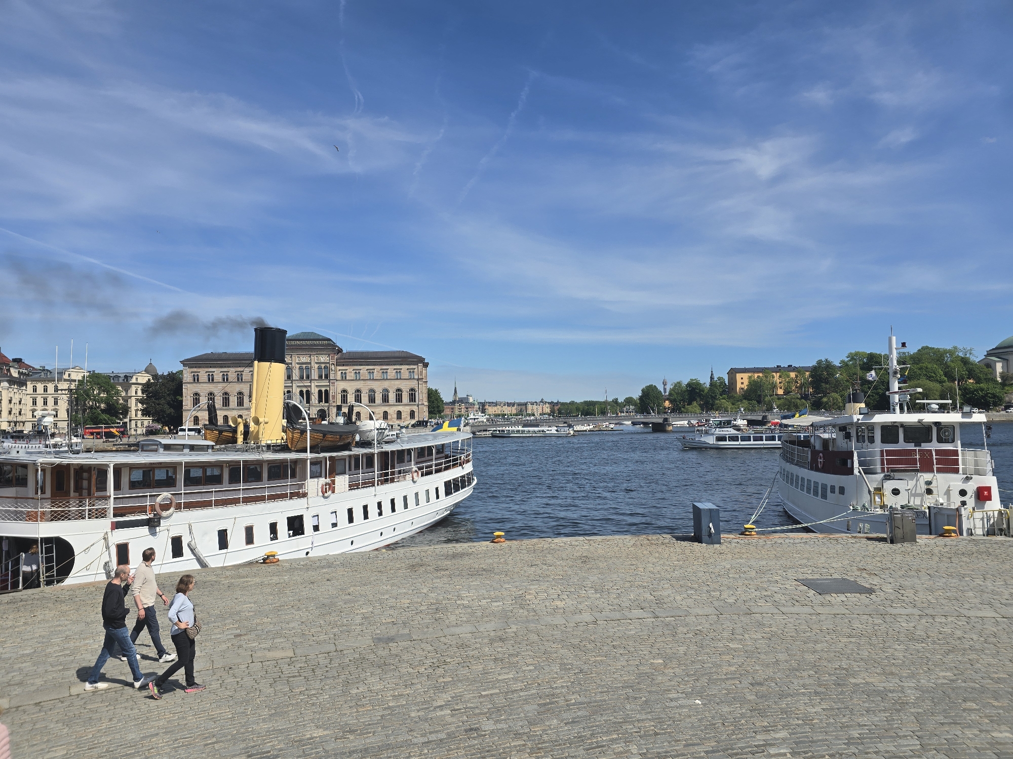 A vibrant daytime scene unfolds on a sunny waterfront, likely in Stockholm, Sweden. In the foreground, a wide cobblestone quay occupies the lower portion of the image. Three individuals are seen walking from left to right across the quay: a man in a black shirt and jeans, another man in a light brown shirt and jeans, and a woman in a light blue top and black pants.
To the left, a large, classic white boat with a tall yellow smokestack is docked, emitting a plume of dark smoke, suggesting it is active or preparing for departure. The boat features wooden lifeboats on its upper deck. On the right side of the frame, a smaller white boat with red accents, bearing the name "REX" faintly on its side, is also moored to the quay.
The water, a broad channel or bay, reflects the bright blue sky, which is dotted with wispy white clouds and a few faint contrails. Across the water, the city skyline is visible, dominated by historical buildings. A prominent structure in the background clearly displays "NATIONALMUSEUM" (National Museum) on its facade. Green trees line the distant shores, and several bridges span the water, with other boats and ferries dotting the waterway. The scene conveys a sense of pleasant activity on a clear, sunny day.