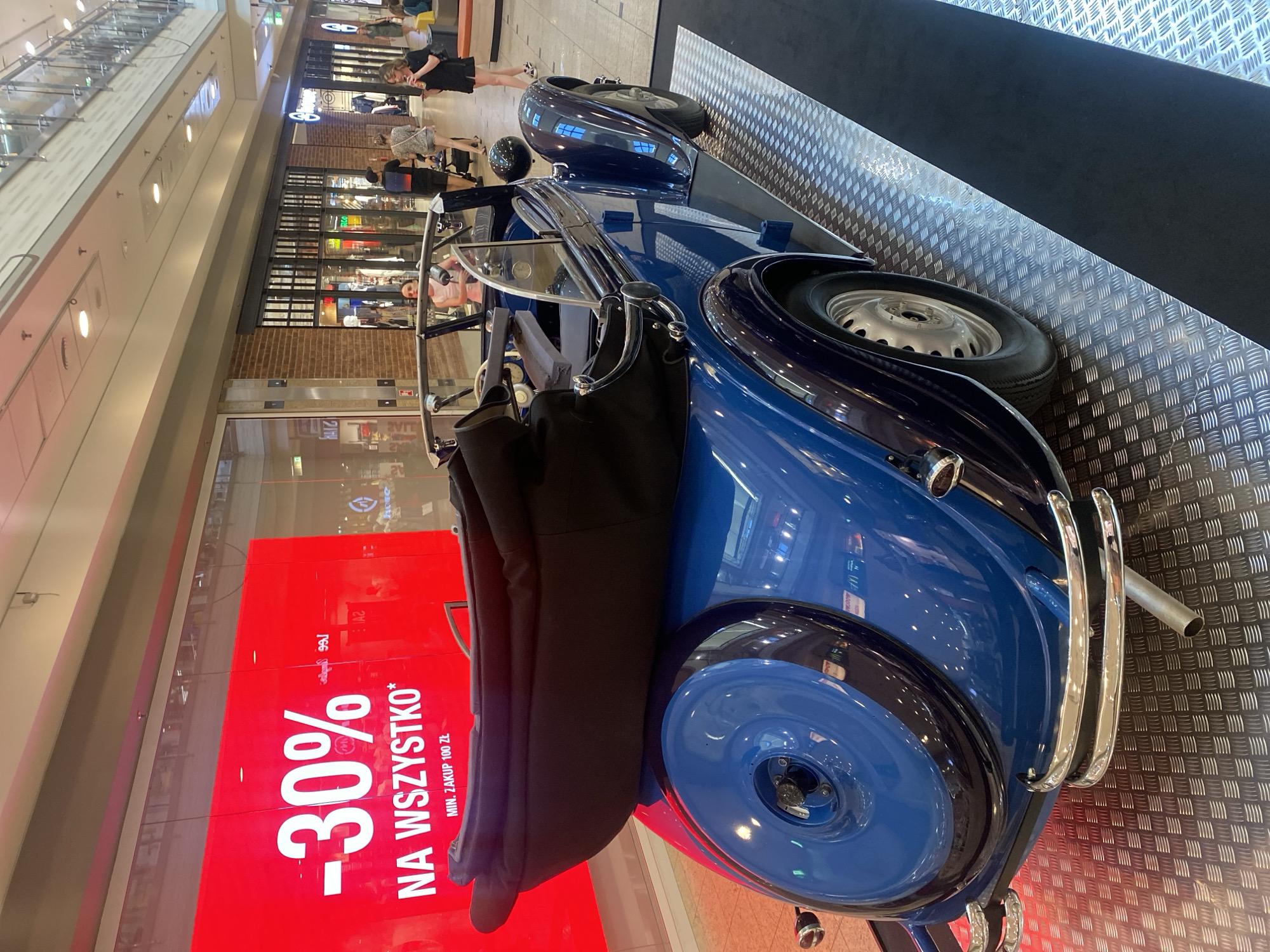 An antique blue convertible car with a black top is on display in a shopping mall. A large red digital screen in the background displays "-30% NA WSZYSTKO* MIN. ZAKUP 100 ZŁ" which translates to "-30% ON EVERYTHING* MIN. PURCHASE 100 PLN". The car is positioned on a metallic checkered floor. In the background, several people are walking and browsing in the mall. The car appears to be a Bugatti Type 57 Atalante. The mall appears to be Manufaktura in Łódź, Poland, judging by the architecture and the presence of brick facades and industrial-style storefronts.