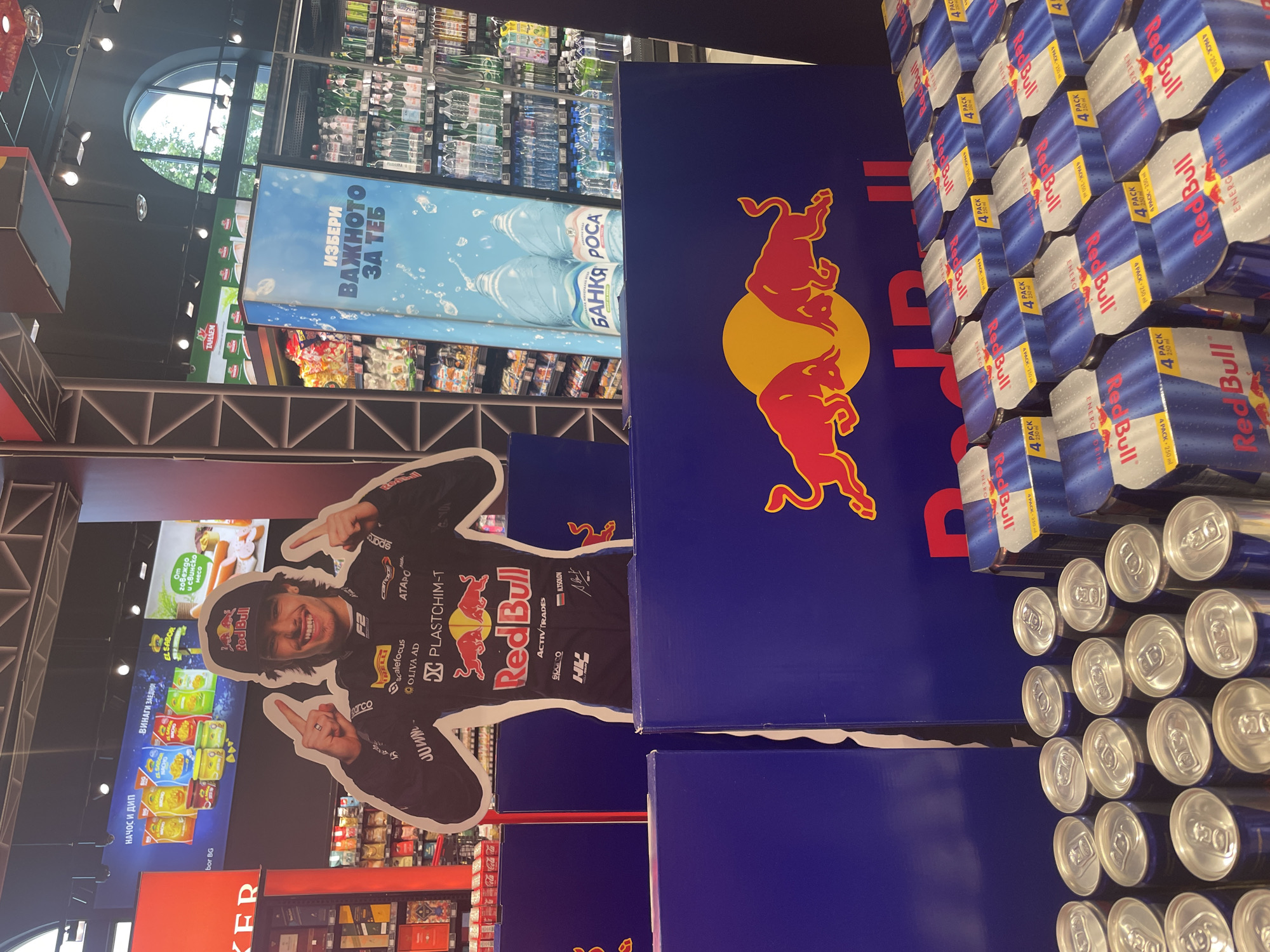 This image shows the interior of a supermarket in Plovdiv, Bulgaria. There is a Red Bull promotional display, including a life-size cutout of a race car driver, and stacks of Red Bull 4-packs. There are various food product advertisements in the background, including chips, meat products, and "Банкя Роса" (Bankya Rosa) bottled water. The scene is well-lit and appears to be a typical retail setting.