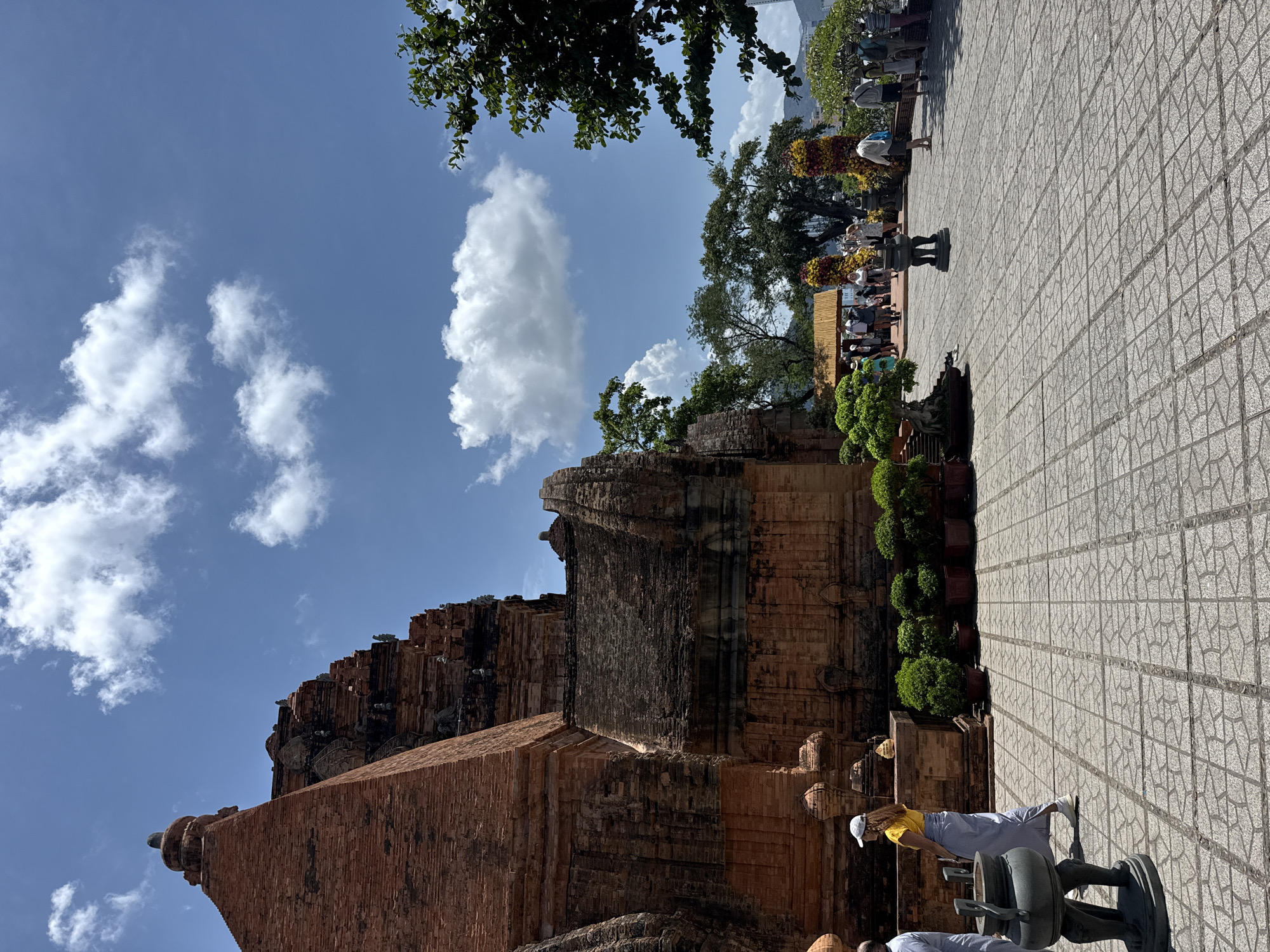 A sideways-oriented, medium-distance, and slightly high-angle shot captures a temple structure in Bac Nha Trang Ward, Vietnam. A blue sky dotted with white clouds forms the backdrop.
The temple is predominantly red-brick, displaying an aged texture. Adjacent to the temple structure, there are numerous potted green plants.
To the right of the image, a paved area is visible with patterned gray tiles. Several people can be seen standing or walking on this paved area. A black metal sculpture stands in the foreground.
The general atmosphere suggests a sunny, outdoor tourist environment.