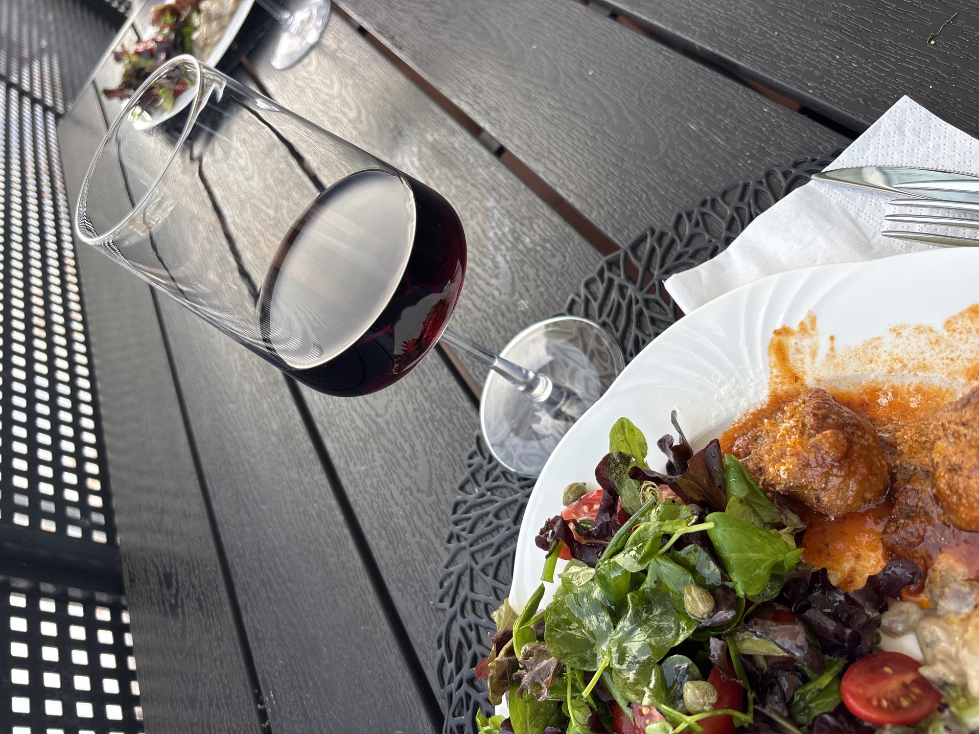 The image depicts a close-up, high-angle view of a meal set on a dark wooden table, likely outdoors given the dappled sunlight and the presence of a latticed structure in the background. A white plate holds a salad with mixed greens, cherry tomatoes, and capers, alongside what appears to be meatballs in a red sauce. A glass of red wine is prominently featured, tilted and reflecting the surrounding light. Cutlery is partially visible, resting on a white napkin. The environment suggests a relaxed dining setting, possibly a restaurant patio or a private outdoor space in Tavira, Portugal. The overall scene captures a moment of dining, with the focus on the food and drink.