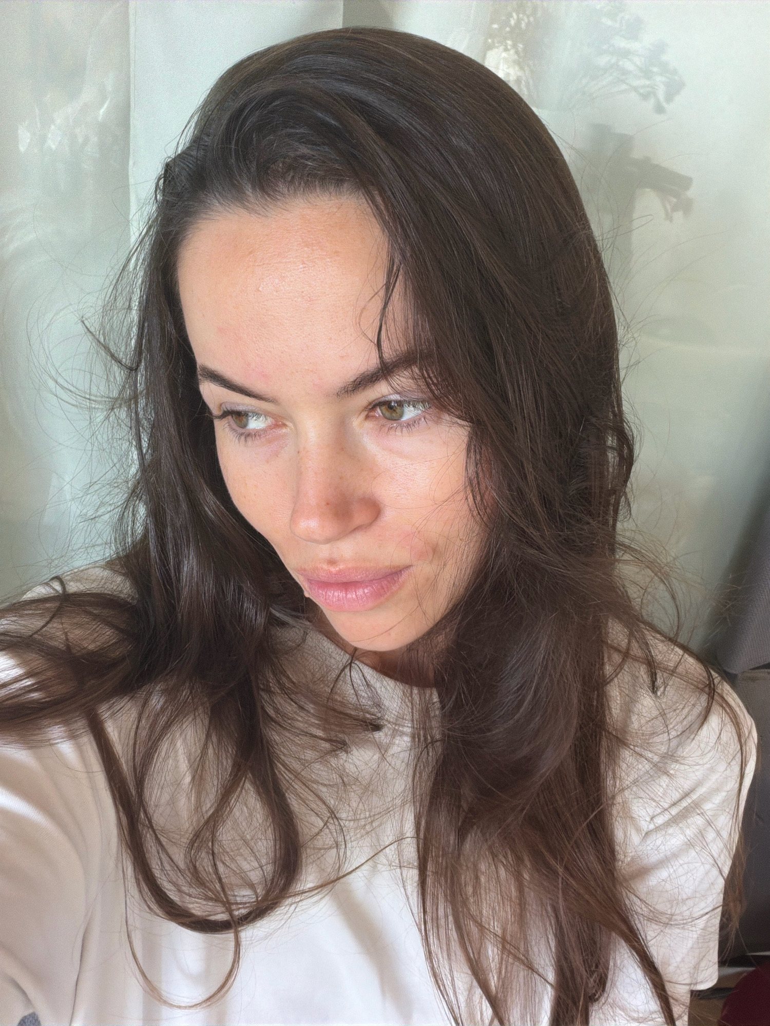 The image is a close-up, waist-up, selfie-style photograph of a young woman with long, dark brown hair. She is looking to her left, with her head tilted slightly downwards. Her skin appears to be light-toned with some subtle redness on her cheeks and forehead, and a few small blemishes are visible. Her lips are slightly parted, and she has light makeup on her eyes. She is wearing a plain white t-shirt. The background consists of a sheer curtain or blinds, behind which shadows of what appear to be plants or flowers are cast. The lighting is soft and diffused, suggesting an indoor setting. There is no discernible indication of the location being Salou, Spain.