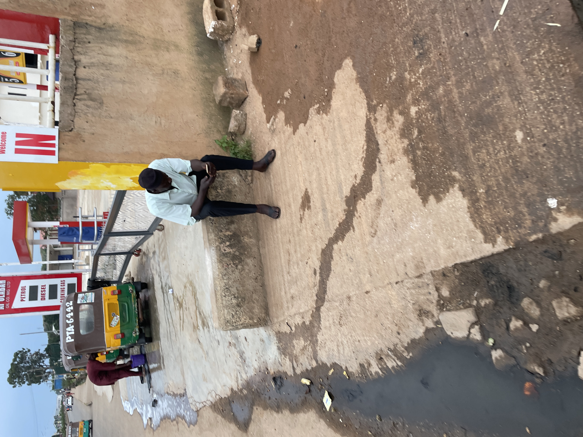 The image shows an outdoor scene at a gas station in Barakin Lerawi, Nigeria. A person is sitting on a concrete block, looking at a mobile phone. They are wearing a light blue shirt and dark pants, with sandals on their feet. Beside them, another person in a maroon outfit is attending to a three-wheeled vehicle, possibly washing it, with a bucket and water nearby. The vehicle has the license plate "PTA-6442". The gas station has prominent signage indicating "PETROL" and "DIESEL". The ground is a mixture of concrete and dirt, with puddles of water and debris. In the background, there are more vehicles, including other three-wheeled vehicles and cars on a road, as well as the structures of the gas station. The sky is overcast.
