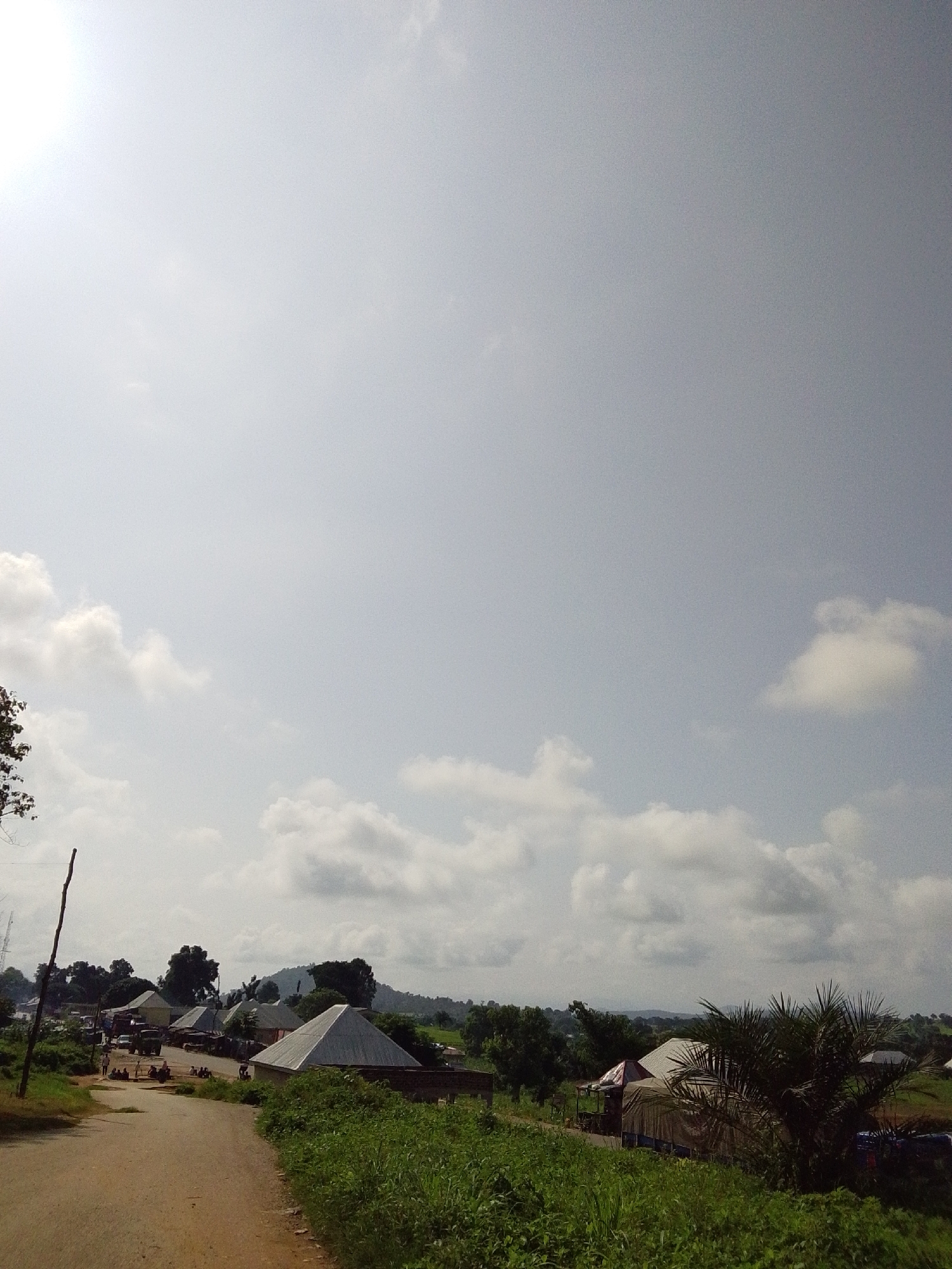 This is a wide shot of a dirt road in the city of Serti, Nigeria, on a bright, cloudy day. The road curves to the left, disappearing into a small village. On the right side of the road, there is lush green grass and foliage. In the distance, there are several buildings with corrugated metal roofs, typical of houses in Nigeria. Some of the buildings appear to be shops or market stalls, with awnings providing shade. There are a few people walking along the road and sitting near the buildings. A vehicle is parked near one of the buildings. In the far distance, there are rolling hills and trees. The sky is bright and overcast with scattered clouds. The scene depicts a typical rural village in Nigeria.