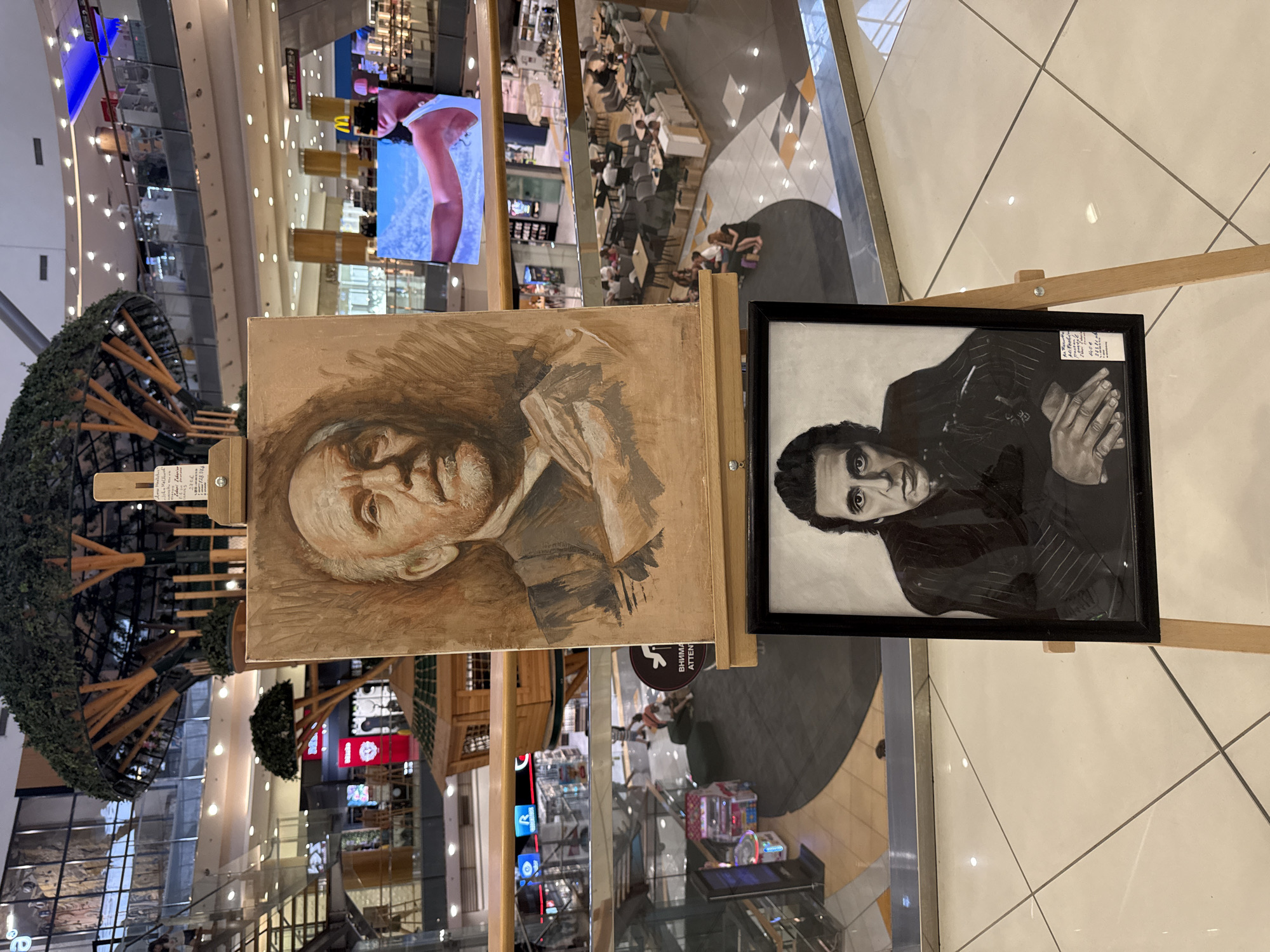 The image shows two framed portraits displayed on wooden easels inside a shopping mall. The upper portrait, on a larger easel, is a painting of an older man's face in a sepia tone, with his hands clasped in front of him. A small sign is attached to the easel, with some text, but it is largely illegible. The lower portrait, framed in black and standing on a smaller easel, is a black and white drawing of a man with dark hair and intense eyes, also with his hands clasped. This easel also has a small sign with text. In the background, the mall environment is visible, with shops, escalators, seating areas with people, and a large screen displaying an image of a person. The floor is tiled. The overall scene suggests an art display or exhibition within a commercial space.