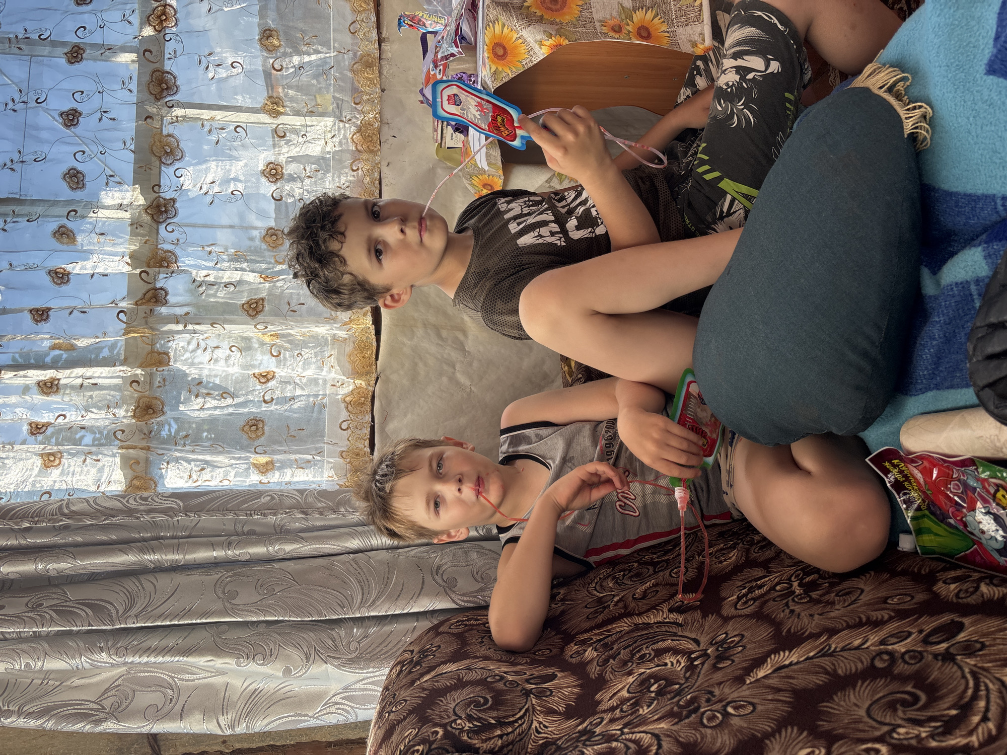 This image captures two young boys indoors, likely in a home in Маркидоновка, Russia. The scene appears to be during the daytime, with sunlight streaming through a patterned sheer curtain on the left. The boys are seated on a patterned sofa.
The boy in the foreground is lying on his side, looking towards the camera. He is wearing a grey tank top with black and red trim and a number "1996" printed on it. He holds a green and pink toy with a tube, possibly for blowing bubbles, in his right hand, with the tube near his mouth.
The older boy is sitting behind and to the right of the foreground boy. He is wearing a dark t-shirt with white lettering and patterned shorts. He is also holding a similar toy with a red tube, with the tube near his mouth, and looking directly at the camera. The toys appear to be bubble wands with connected pouches.
In the background, a wooden table is visible with a floral tablecloth and some items on it, including what looks like packaged drinks or snacks. The overall atmosphere seems relaxed and casual, suggesting a moment of playtime or leisure. The sunlight creates a warm glow in the room, and the boys' expressions are neutral.